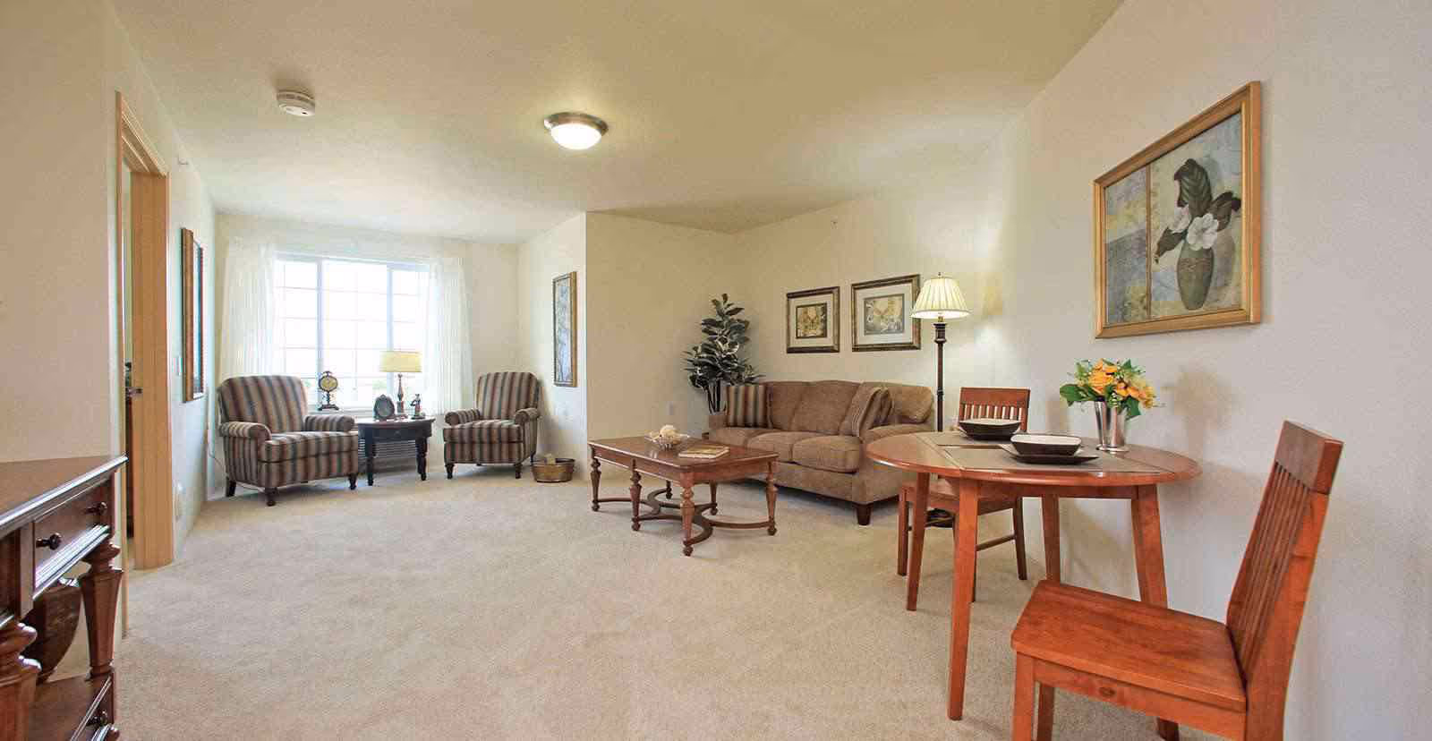 A bright living room with beige carpet and walls, featuring a brown sofa with two cushions, a wooden coffee table, two striped armchairs near a large window with white curtains, a small wooden dining table with two chairs, a floor lamp, framed artwork on the walls, and a potted plant in the corner.