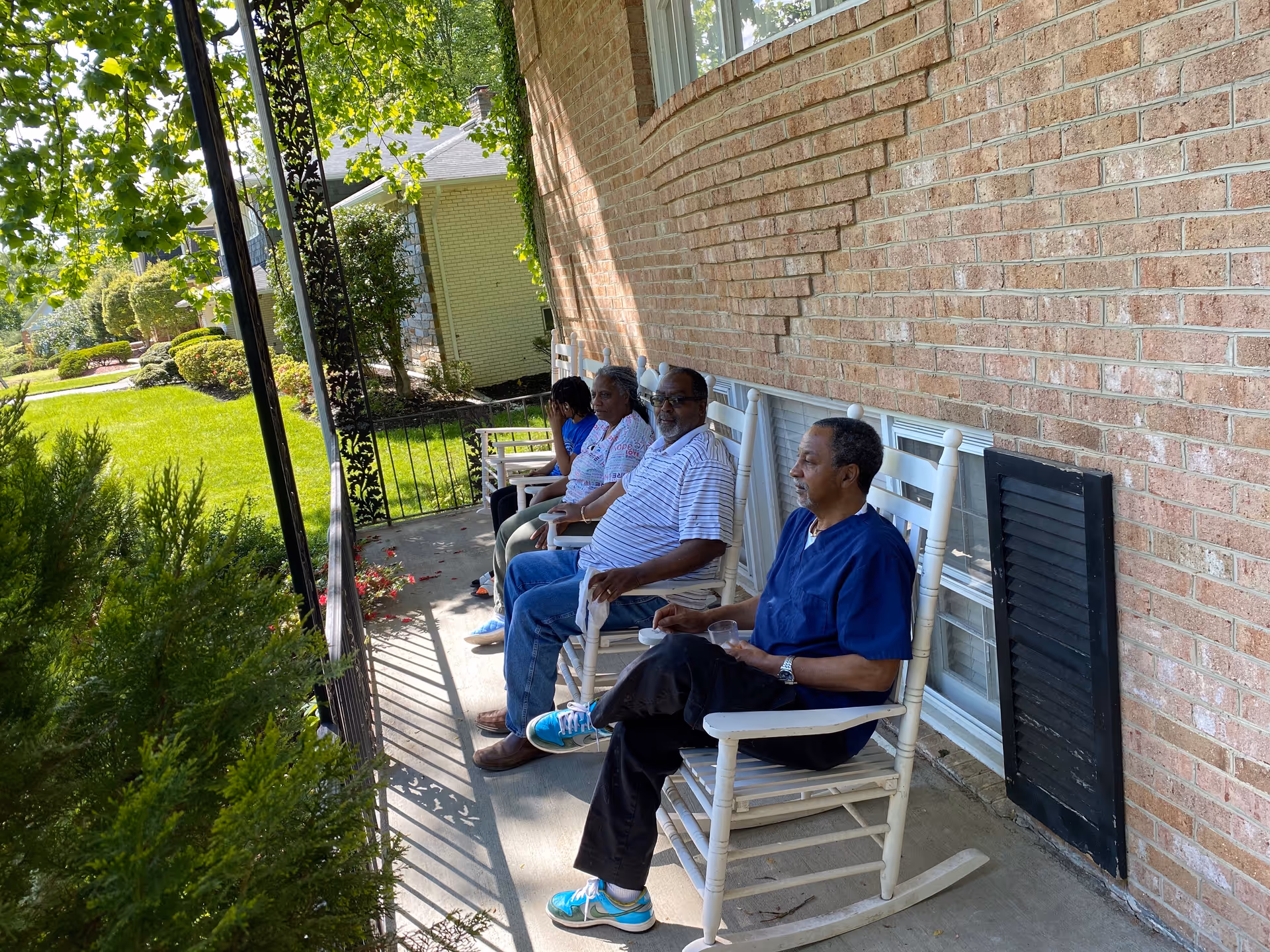 Four people sitting on white rocking chairs on a porch next to a brick wall, with greenery and trees visible in the background on a sunny day.