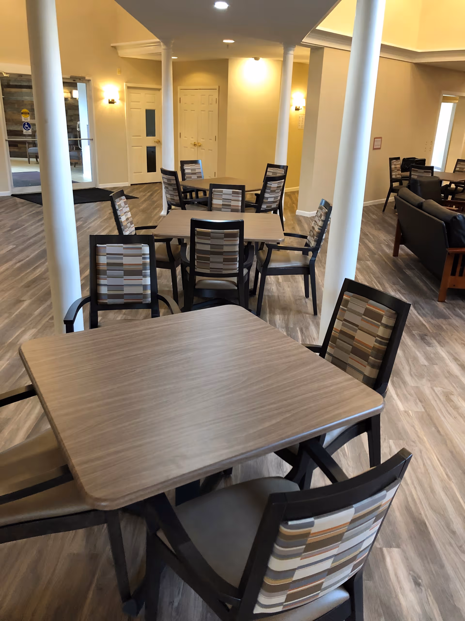 Interior view of a senior living facility common area with multiple square tables surrounded by chairs with patterned backs. The room has light-colored walls, wood flooring, white columns, and soft lighting. There is a seating area with a couch visible in the background.