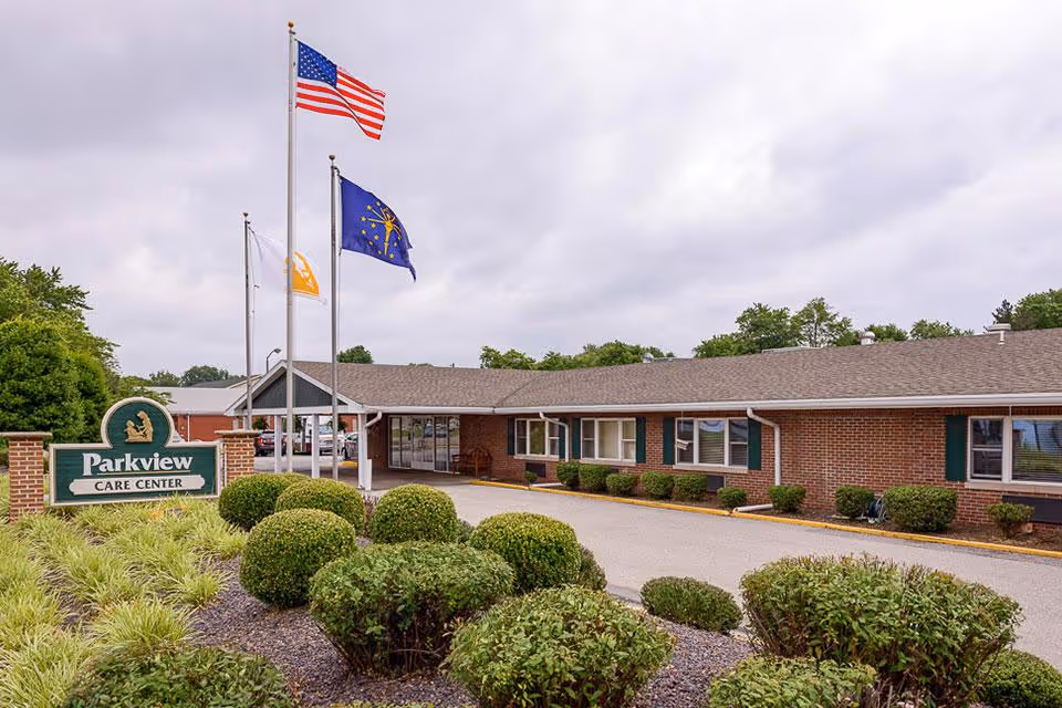 Exterior view of Parkview Care Center, a single-story brick building with green shutters and a covered entrance. In front of the building are neatly trimmed bushes and a sign that reads 'Parkview Care Center'. Three flagpoles display the American flag, a state flag, and another flag. The sky is overcast.