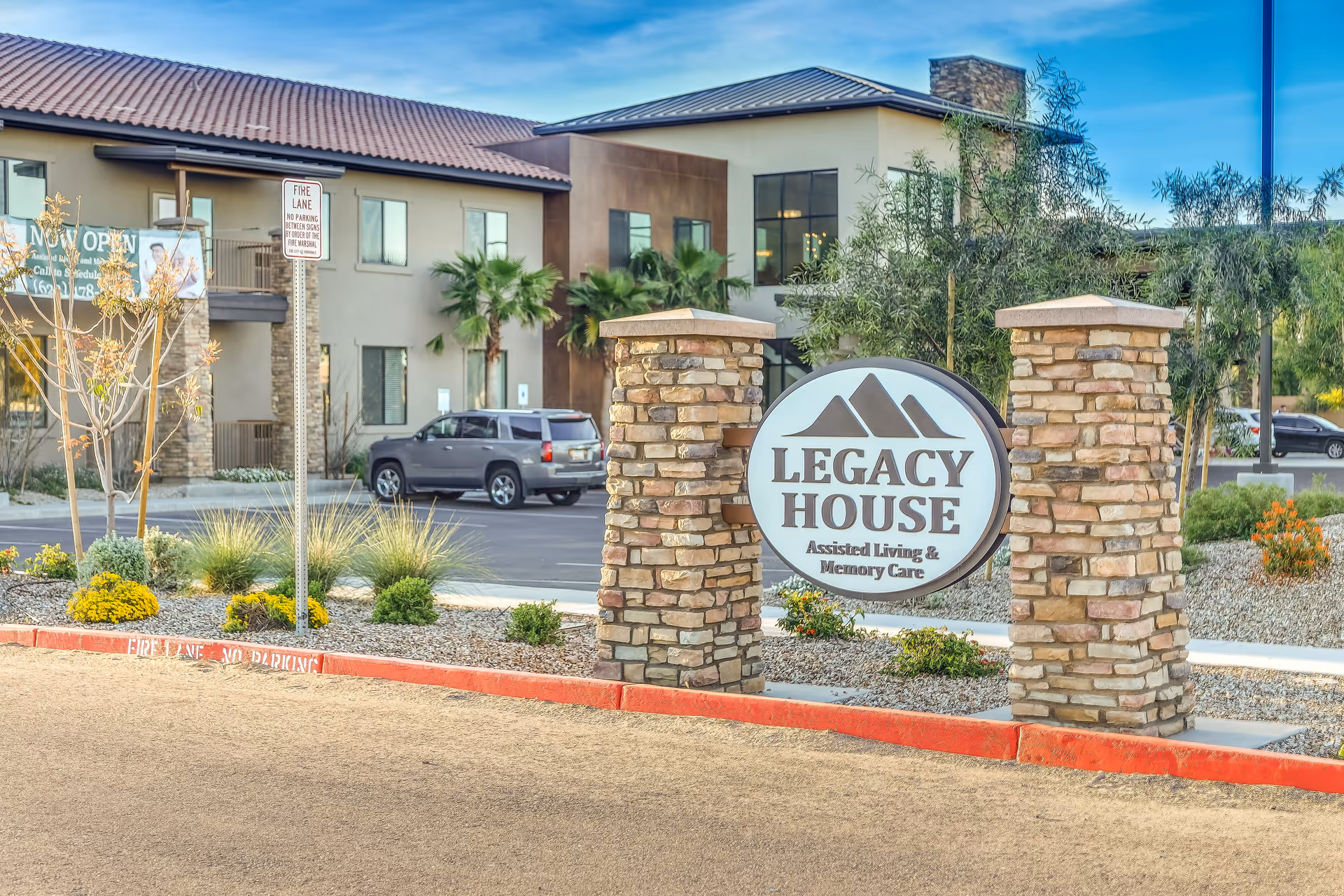 Exterior view of Legacy House Assisted Living & Memory Care facility with a stone sign in the foreground and a two-story building with parked cars in the background under a blue sky.