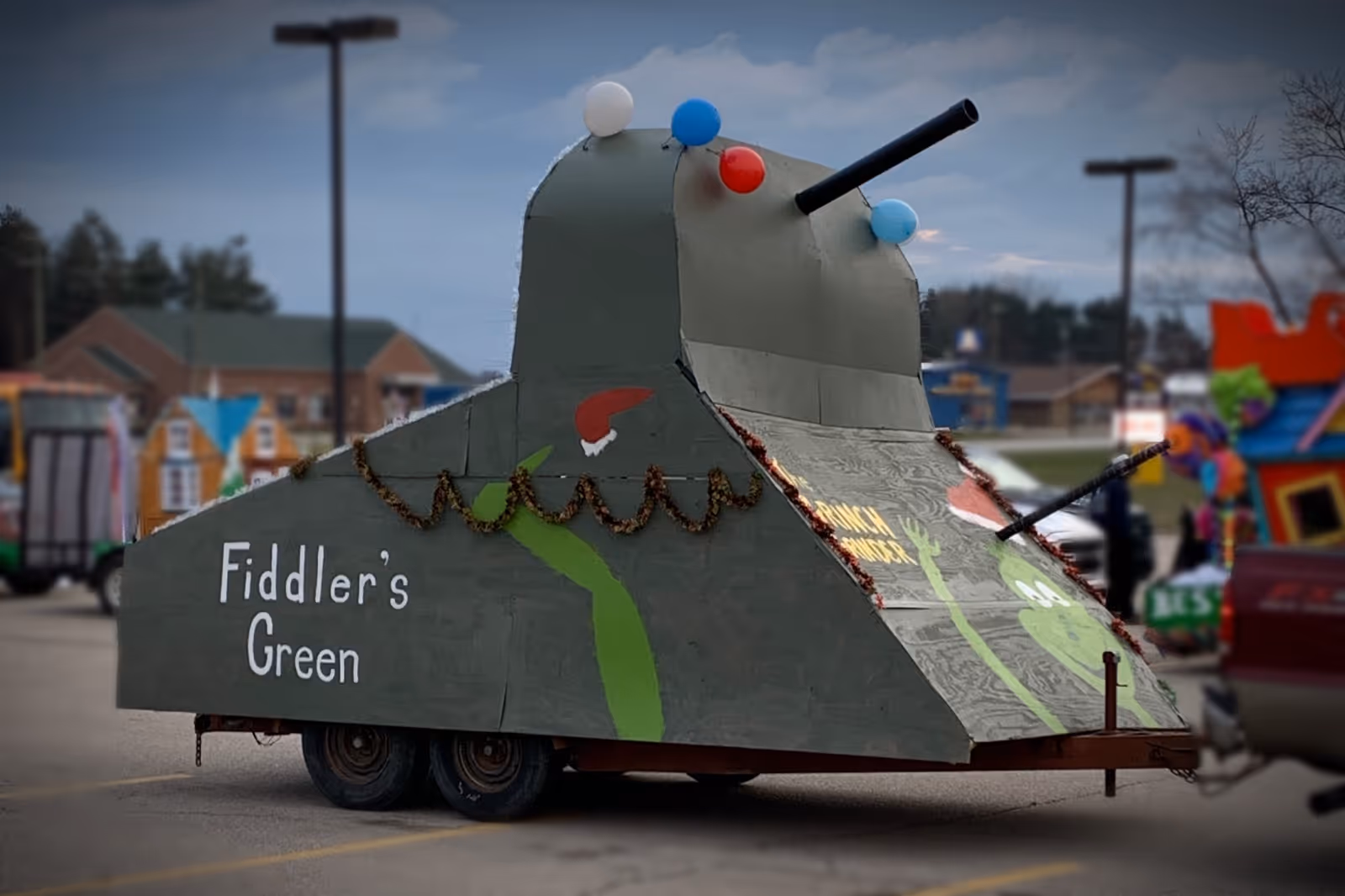 A green tank-shaped parade float on a trailer with 'Fiddler's Green' painted on the side and holiday decorations in a parking lot.