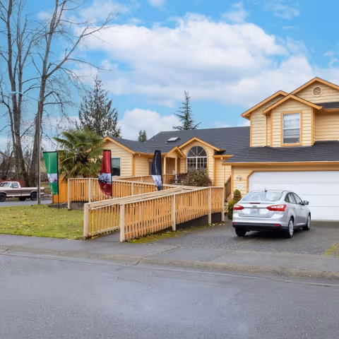 Exterior view of a two-story yellow house with a wooden ramp leading to the front entrance. A silver car is parked in the driveway next to a white garage door. The sky is partly cloudy and there are trees and grass around the house.