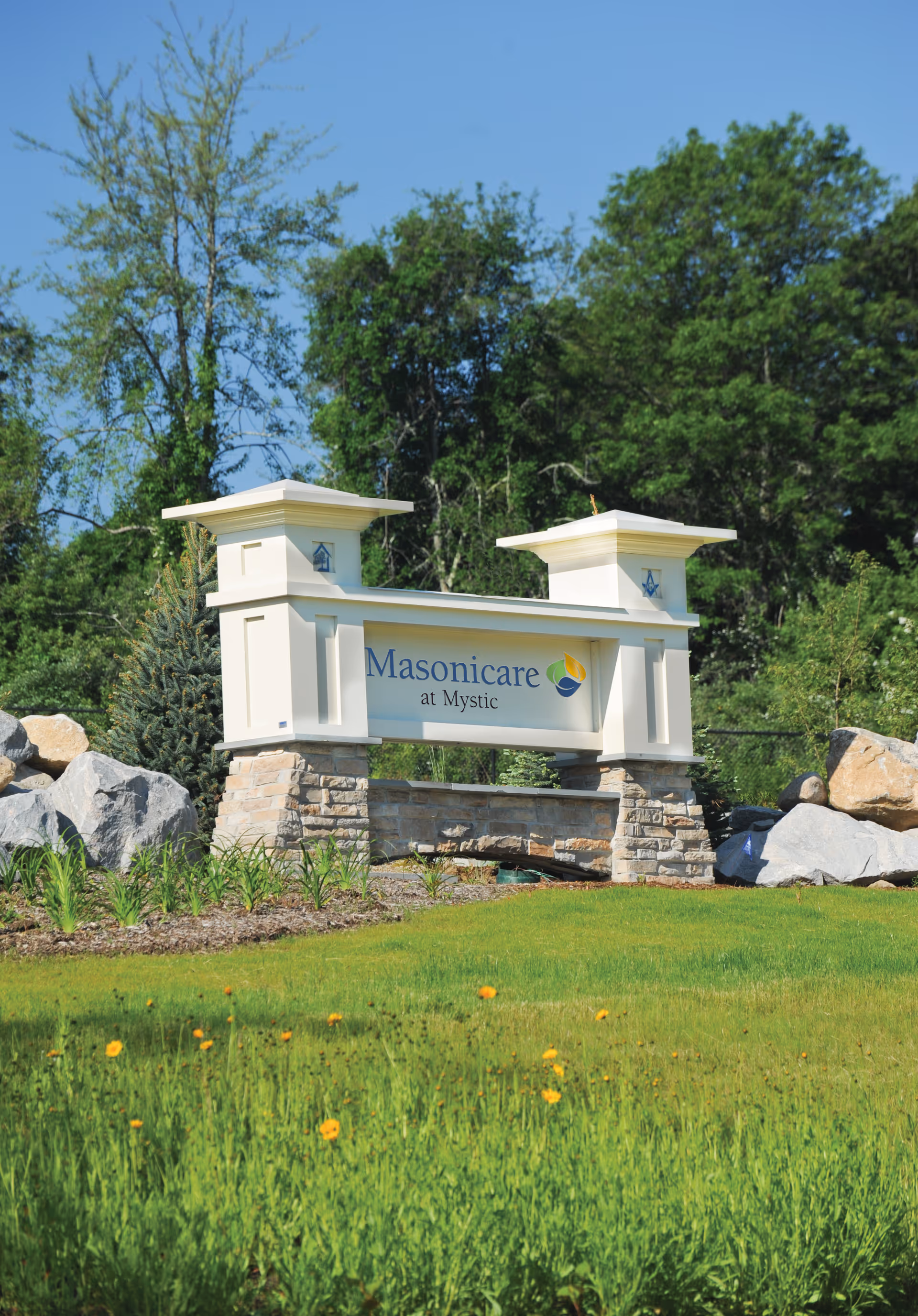 An entry sign reading "Masonicare at Mystic" mounted on stone pillars in a grassy landscaped area with trees in the background.