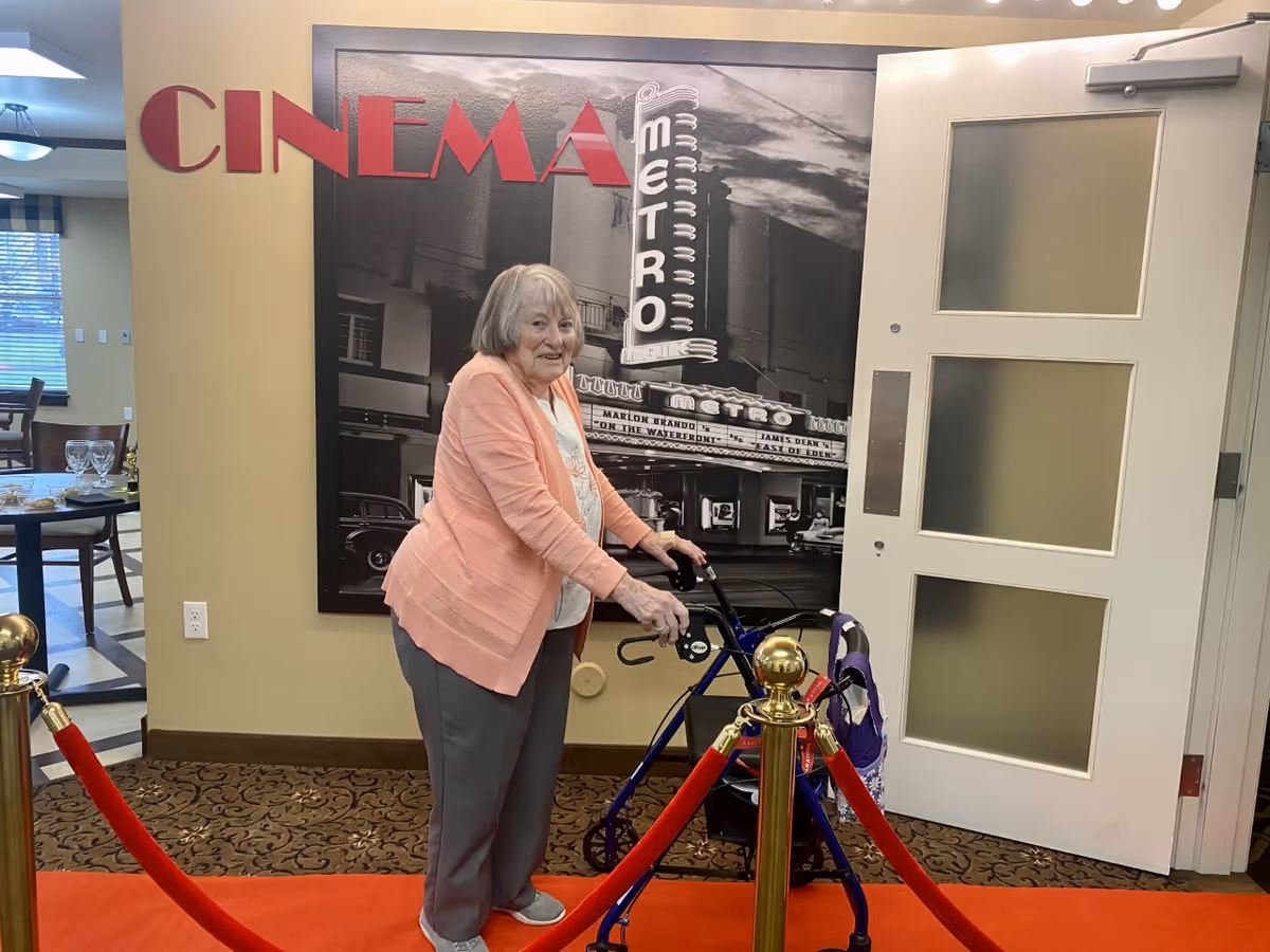 An elderly woman with gray hair using a walker stands on a red carpet in front of a wall with a large black and white photo of a vintage cinema marquee that reads 'METRO'. The word 'CINEMA' is displayed in large red letters on the wall. The area is roped off with gold posts and red velvet ropes. To the left, there is a dining area with tables and chairs visible through an open doorway.