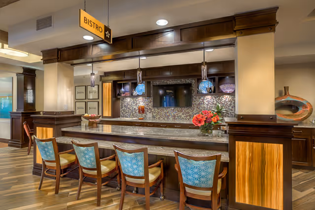 Interior view of a bistro area in an assisted living facility featuring a long counter with five wooden chairs that have blue patterned backs and beige seats. Above the counter are three pendant lights with decorative shades, and a sign hanging from the ceiling reads 'BISTRO'. The back wall has a mosaic tile backsplash, a mounted flat-screen TV, and various decorative items including a vase with red flowers on the counter.