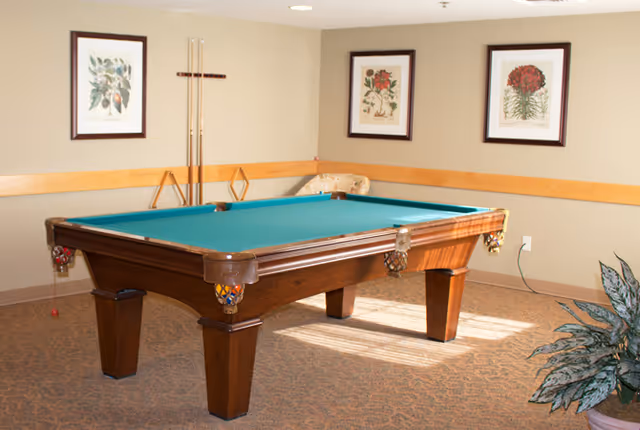 A billiards room with a green felt pool table in the center, three framed botanical prints on the beige walls, a set of pool cues mounted on the wall, and a potted plant in the corner.