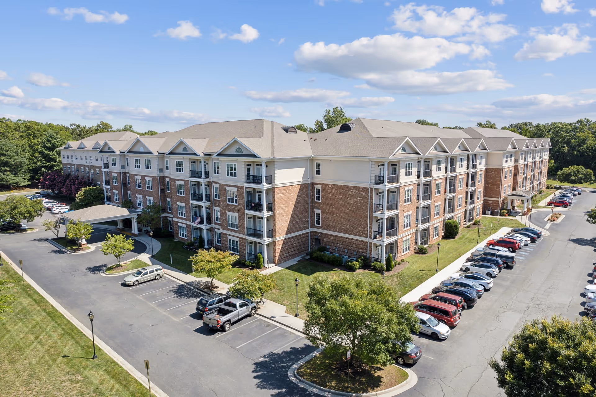 A large multi-story senior living facility building with brick and light-colored exterior walls, surrounded by parking lots with numerous parked cars and some trees. The sky is partly cloudy with blue patches.