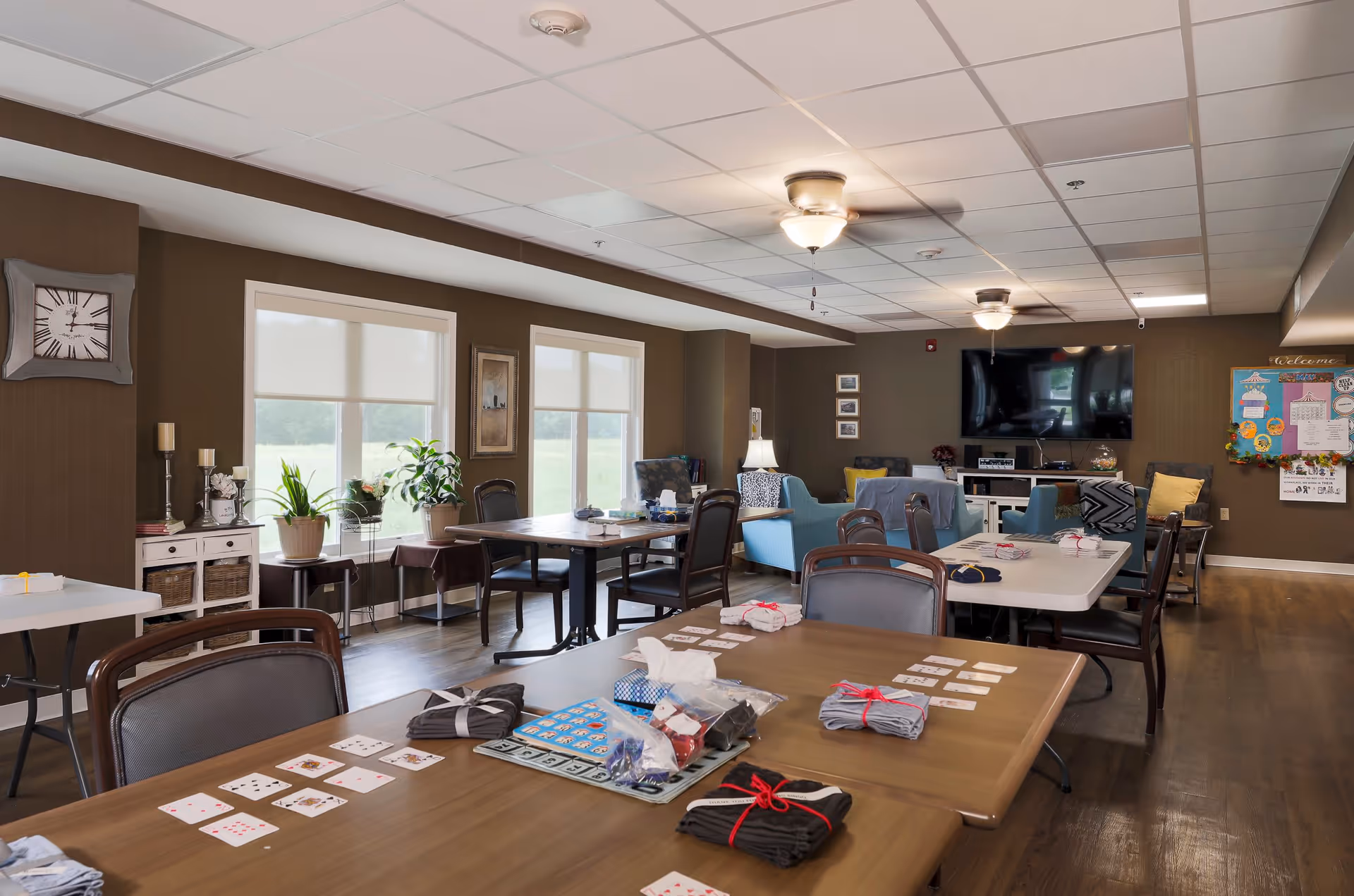 A well-lit common area in an assisted living facility with several tables and chairs arranged for group activities. Playing cards and game materials are spread out on the tables. The room has large windows with blinds, potted plants, a wall clock, and comfortable seating including blue armchairs. A large flat-screen TV is mounted on the far wall above a white cabinet, and a bulletin board with colorful notices is visible on the right side.
