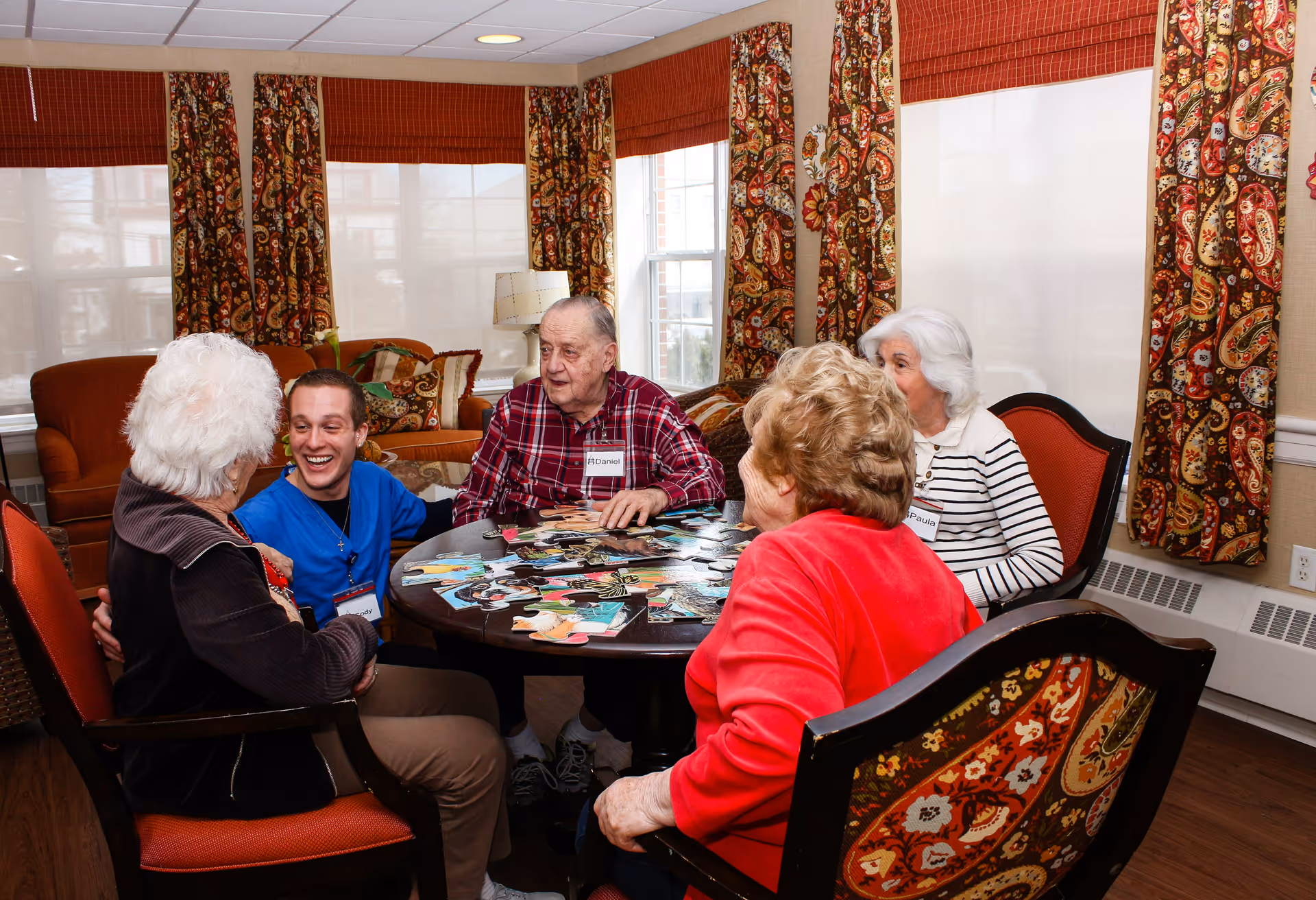 Several older adults and a younger caregiver sit around a table in a bright living room working on a jigsaw puzzle.