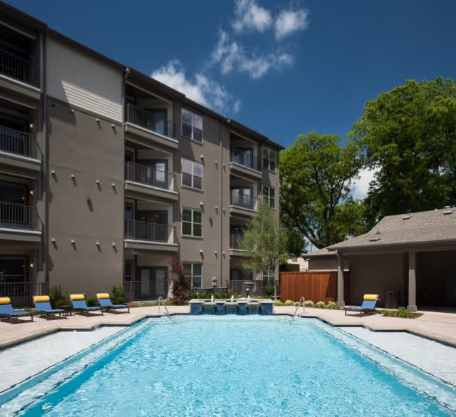 Outdoor swimming pool with clear blue water surrounded by lounge chairs with yellow cushions, adjacent to a multi-story residential building and a small covered structure, under a partly cloudy blue sky.