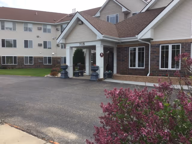 Covered front entrance of a senior living building marked 715 with planters and flowering shrubs in the foreground.