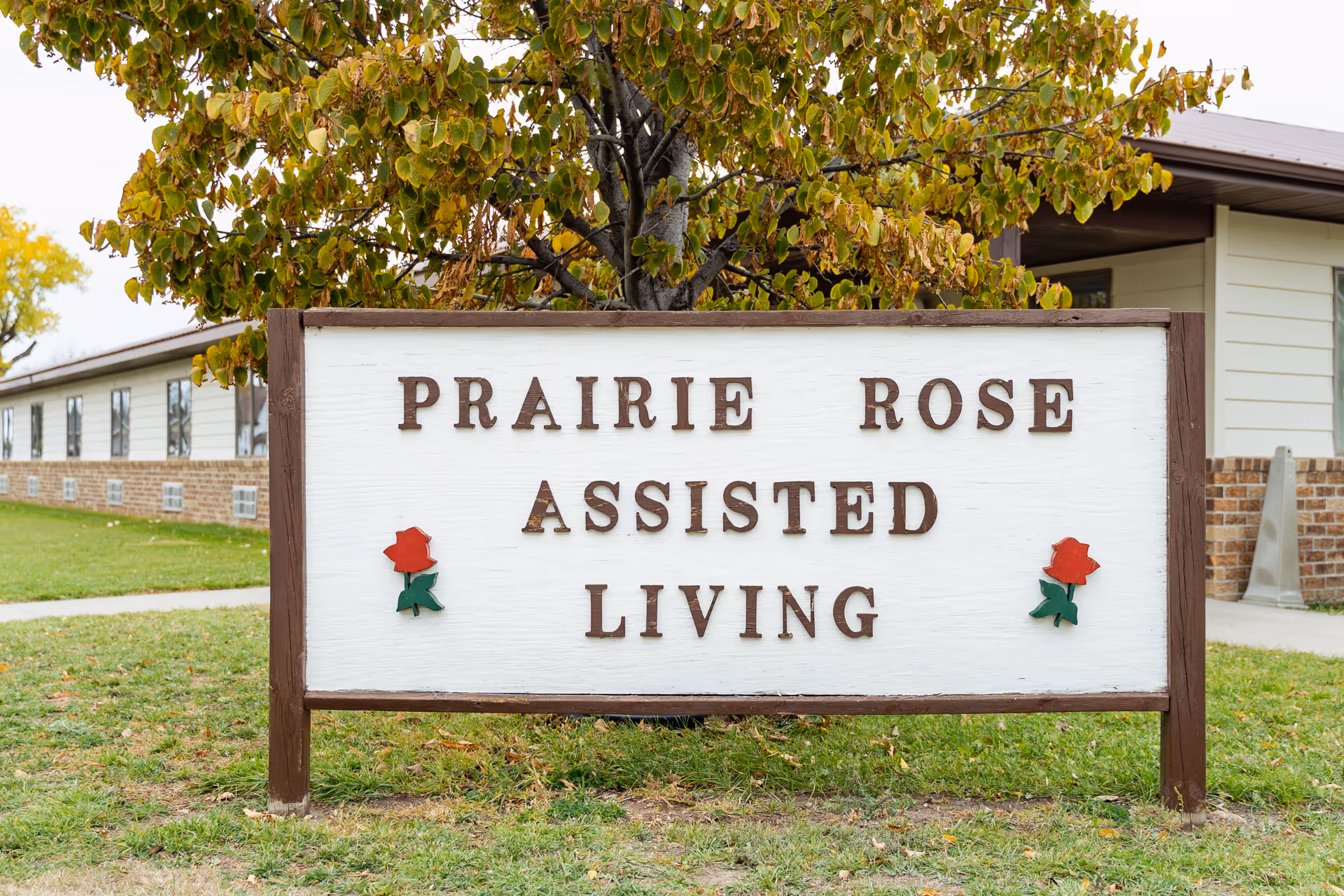 A wooden sign that reads 'PRAIRIE ROSE ASSISTED LIVING' set on grass in front of the facility with a tree and building behind.