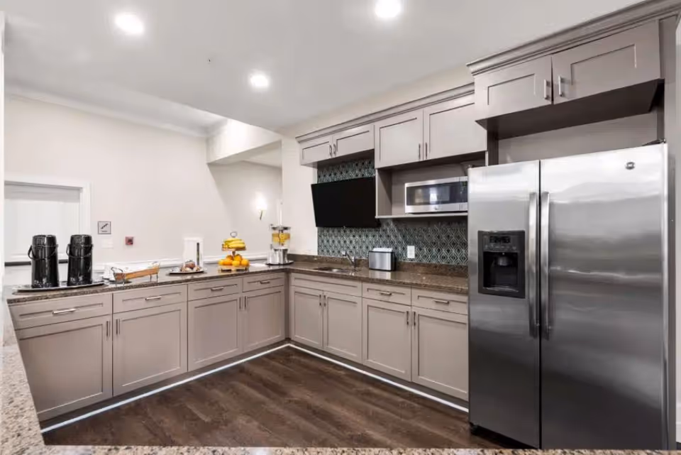 Modern kitchen area with light gray cabinets, a stainless steel refrigerator, microwave, and coffee station on the countertop. The backsplash features a patterned tile design, and the floor is dark wood.