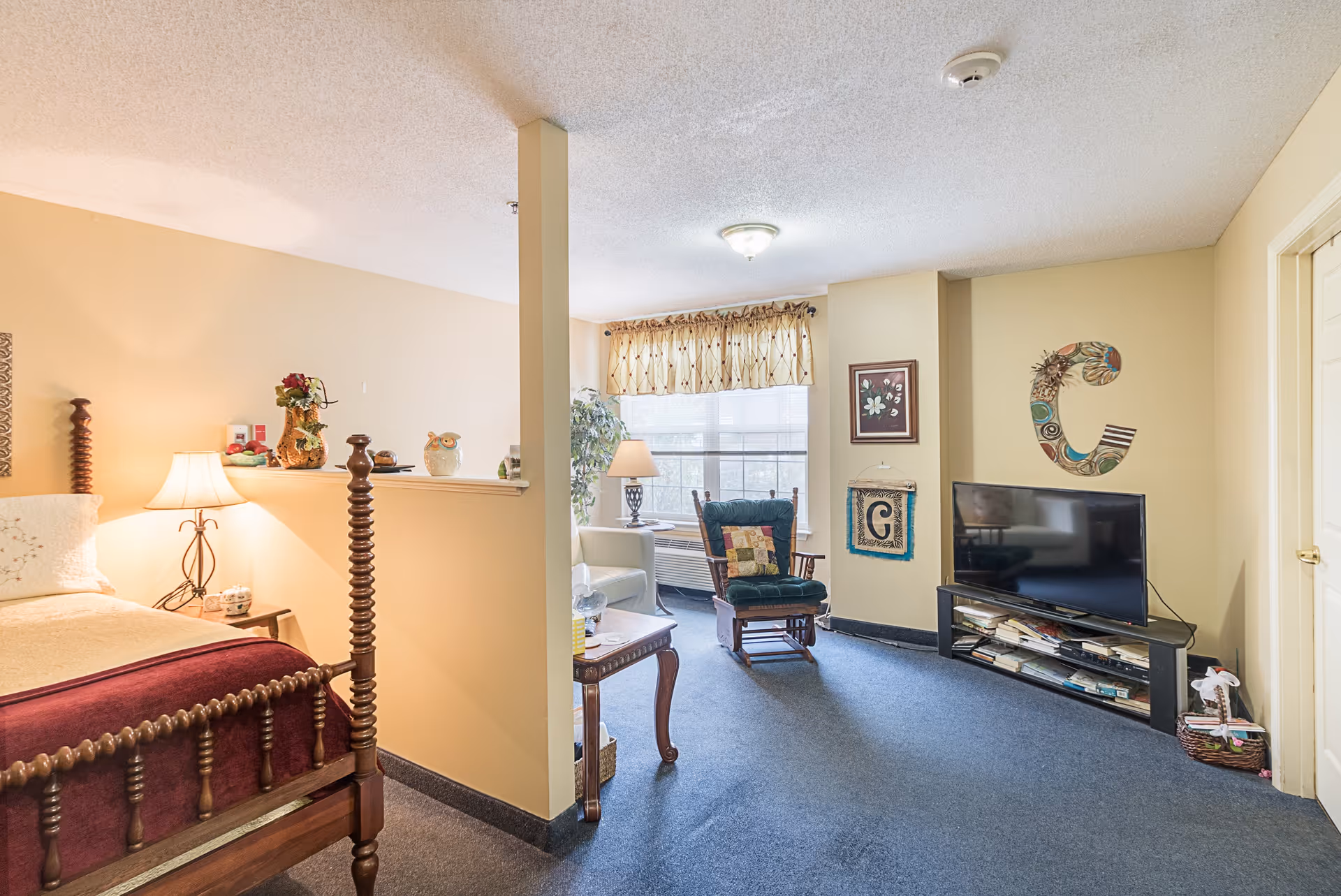 Studio-style assisted living suite with a bed, partial divider, rocking chair by a window, TV on a stand, and wall decorations.