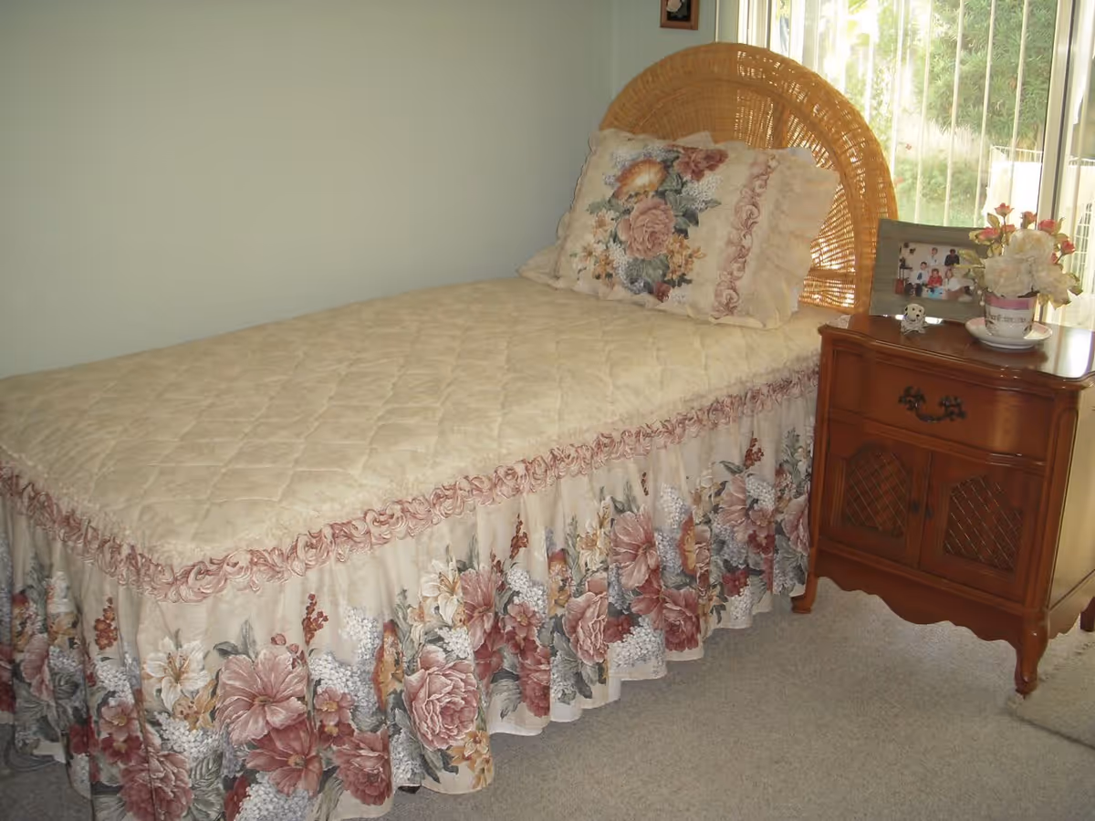 A single bed with a floral bedspread and matching pillow in a room. Next to the bed is a wooden nightstand with a framed photo, a small decorative owl, and a vase with flowers. A window with vertical blinds is visible in the background.