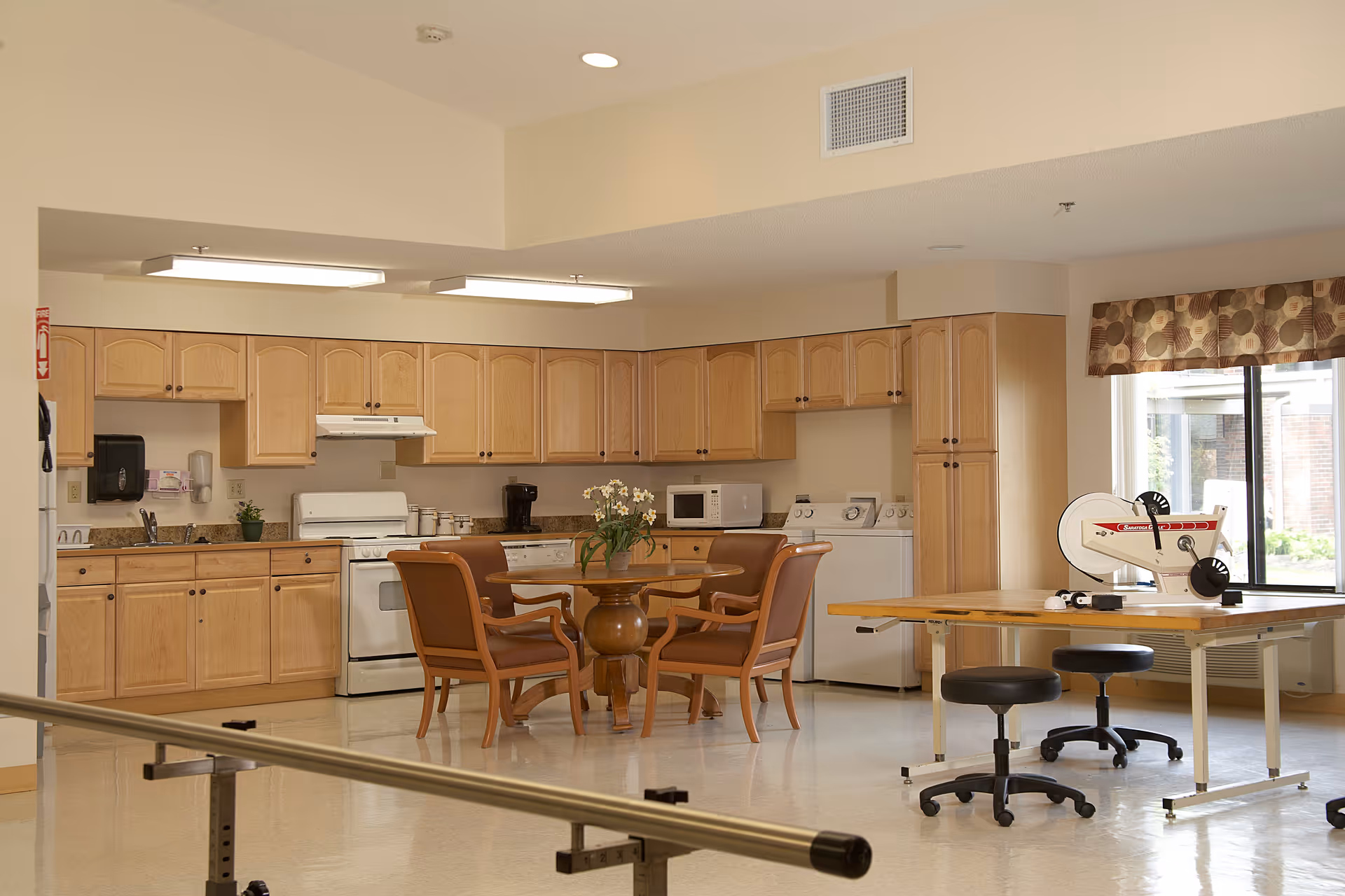 A spacious room with a kitchen area featuring light wood cabinets, a white stove, microwave, and dishwasher. In front of the kitchen is a round wooden table with four brown cushioned chairs. To the right, there is a therapy or exercise table with two black stools and a piece of exercise equipment. A large window with patterned valance lets in natural light.