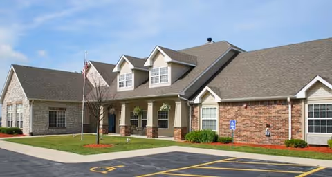 Single-story brick and stone senior living facility front with a covered porch, dormer windows, flagpole, and accessible parking.