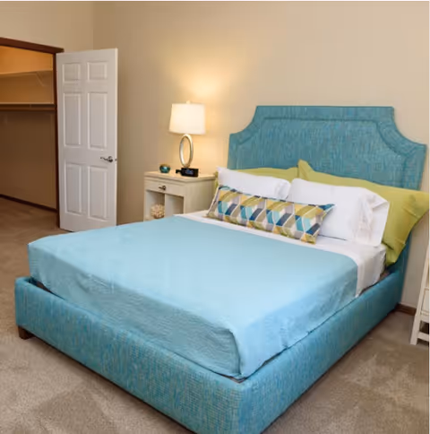 A bedroom with a teal upholstered bed and matching bedspread, a nightstand with a lamp, and an open closet door.
