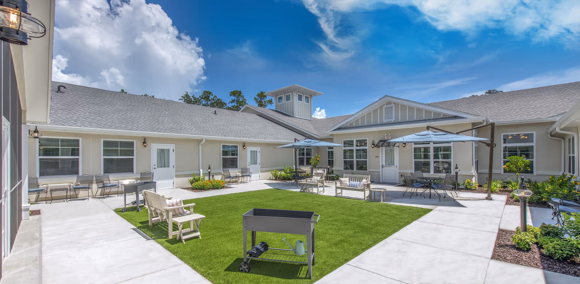 Outdoor courtyard area at The Addison of Oviedo featuring a green lawn surrounded by concrete walkways, patio furniture including benches, chairs, tables with umbrellas, and a barbecue grill. The building exterior is beige with multiple windows and doors under a clear blue sky with some clouds.