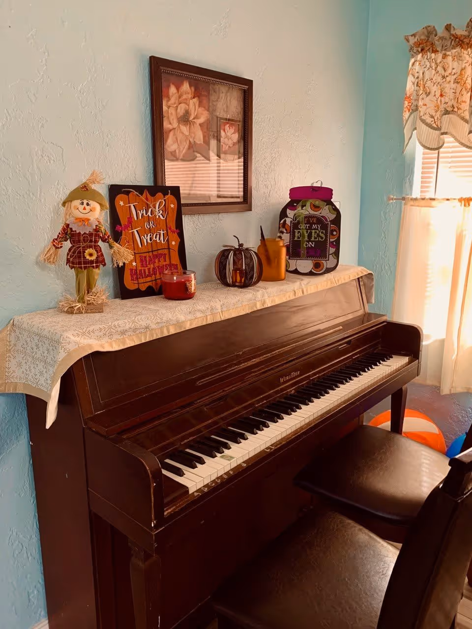 An upright piano with a lace runner on top, decorated with Halloween-themed items including a scarecrow doll, a 'Trick or Treat' sign, a pumpkin candle holder, and a jar with a purple lid. A framed floral picture hangs on the light blue wall above the piano. Two black piano benches are in front of the piano, and a window with floral curtains and white blinds is to the right.