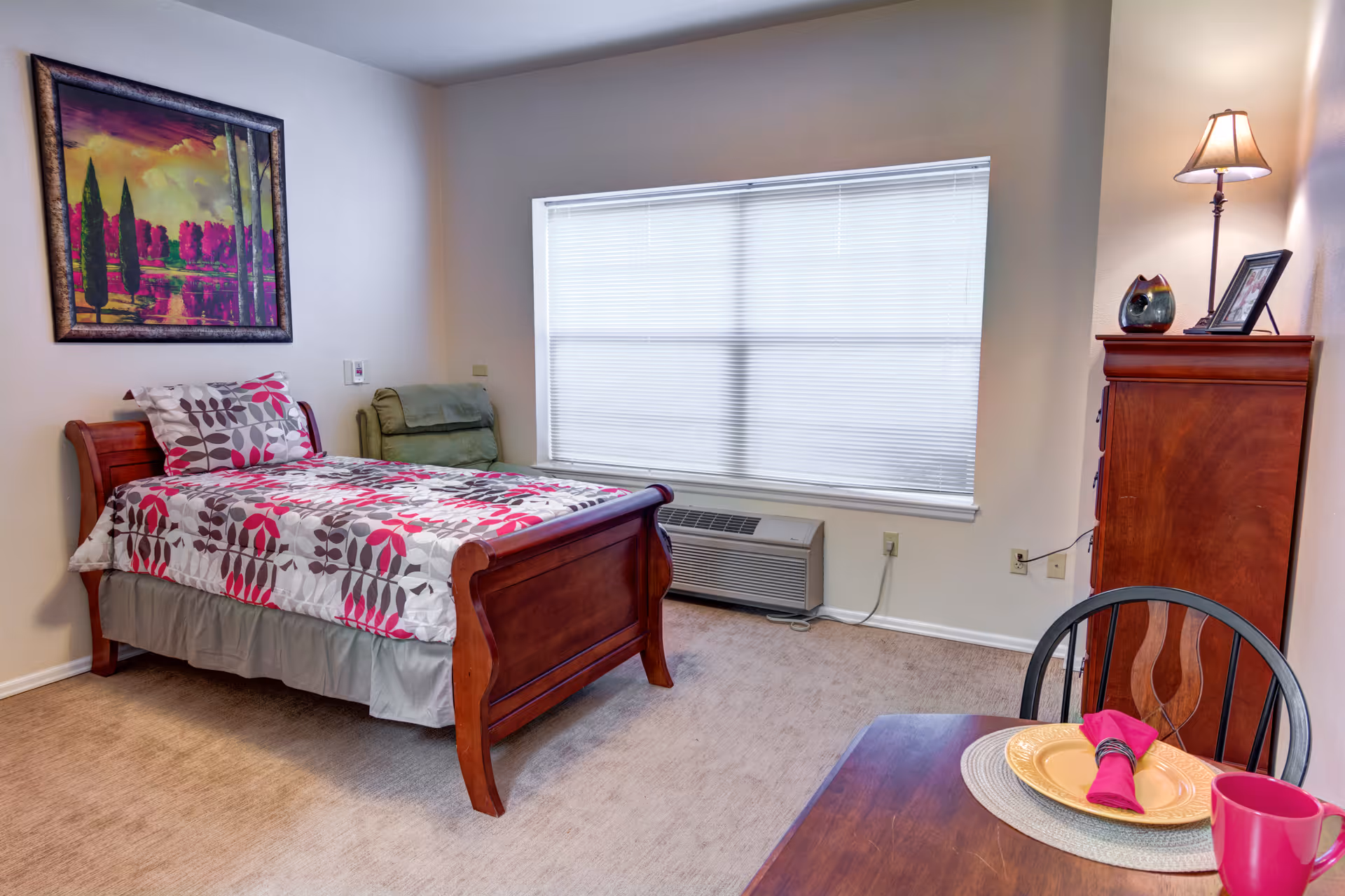 Sunlit furnished bedroom with a wooden twin bed with floral bedding, a dresser, armchair and small dining table by a large window.
