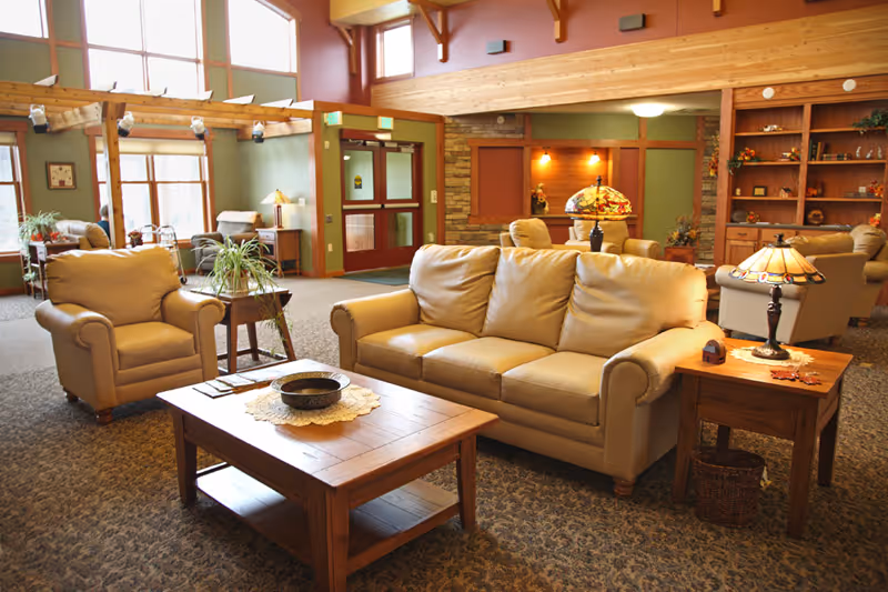 A spacious living room area with beige leather sofas and armchairs arranged around wooden coffee and side tables. The room features large windows letting in natural light, wooden beams, built-in shelves with decorative items, and warm lighting from table lamps.