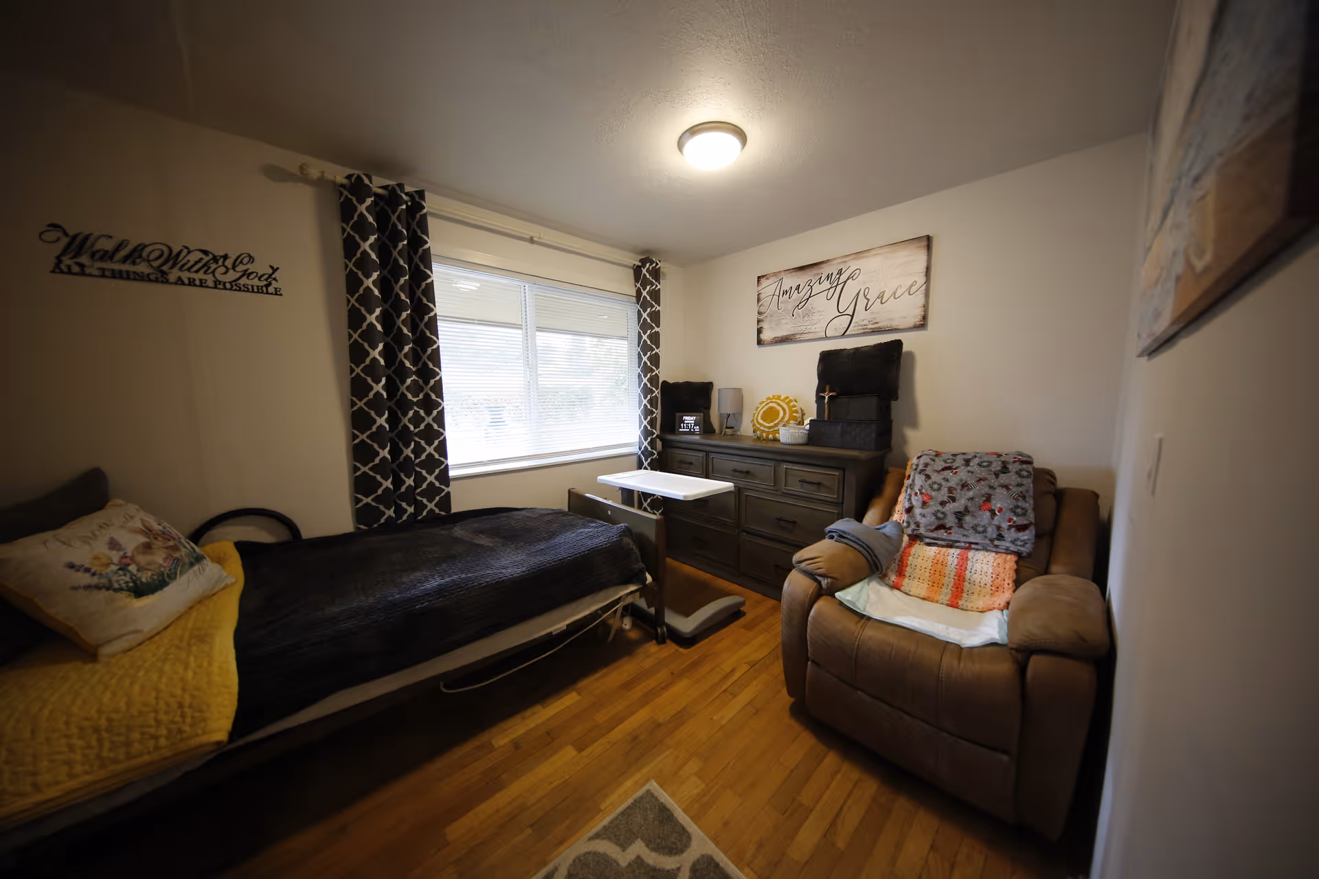 Small furnished bedroom with a single bed, overbed table, recliner chair, dresser under a window and wall decor reading 'Amazing Grace'.