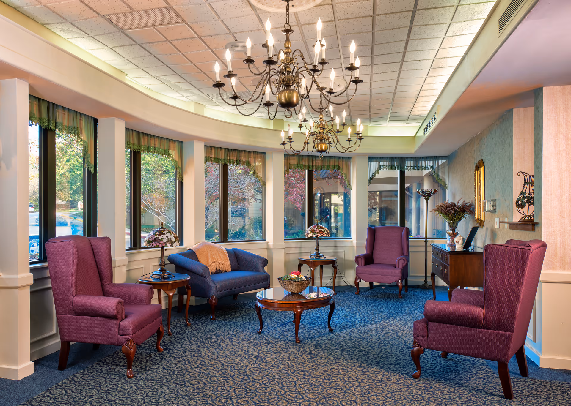 A cozy sitting area with four purple armchairs and a blue loveseat arranged around a wooden coffee table and side tables. The room has large windows with green valances, blue patterned carpet, and two ornate chandeliers hanging from the ceiling. There is a wooden sideboard with decorative items and a mirror on the wall.