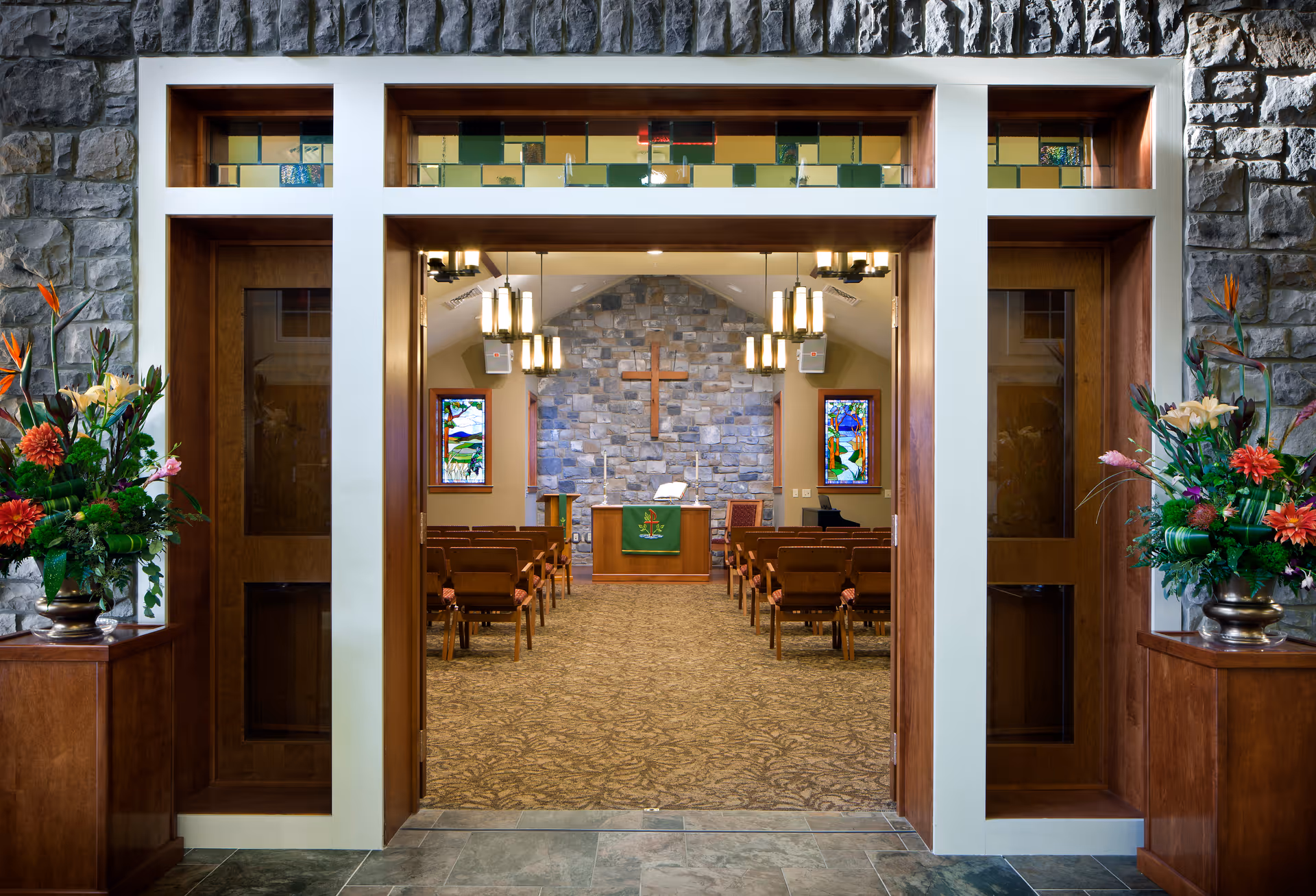 View through the entrance of a chapel with wooden doors and stone walls. Inside, there are rows of wooden chairs facing an altar with a green cloth, a wooden cross mounted on a stone wall, and two stained glass windows on either side. Floral arrangements are placed on wooden stands outside the entrance.