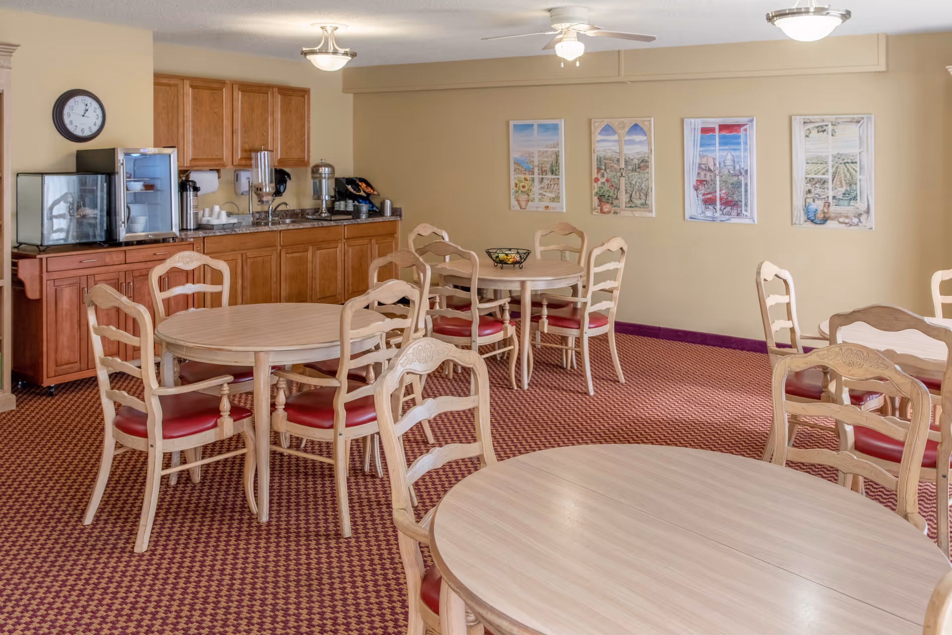 A dining room with several round wooden tables and chairs with red cushions. The room has a patterned carpet and beige walls decorated with framed pictures. There is a kitchenette area with wooden cabinets, a small refrigerator, a coffee maker, and a clock on the wall.