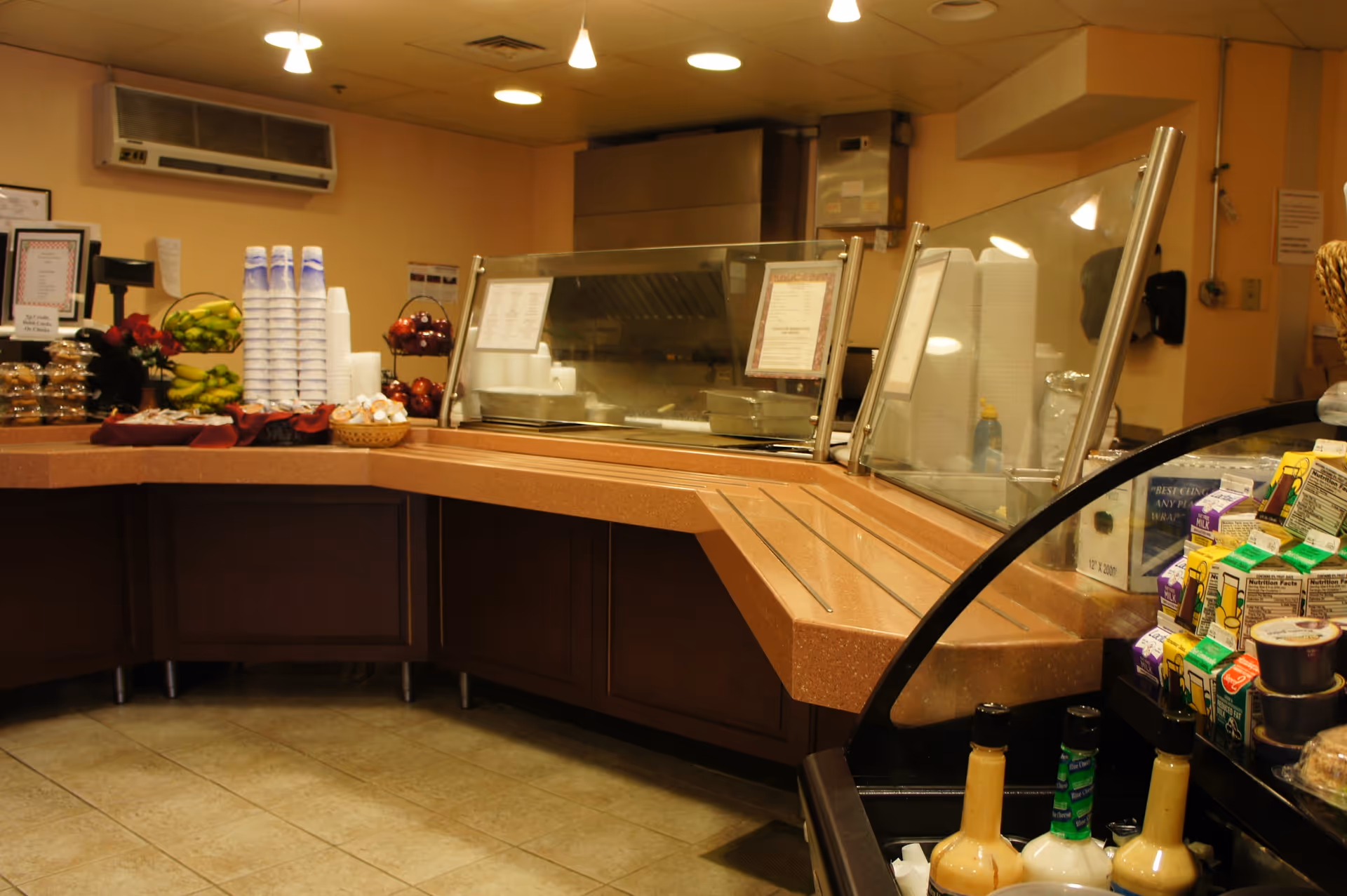 Cafeteria-style serving counter with a glass sneeze guard, stacked cups, fruit and beverage cartons in a dining area.
