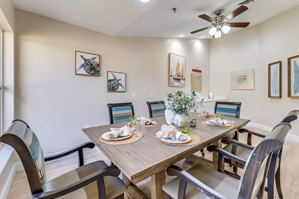 Well-lit dining room with a wooden table set for six, upholstered chairs, wall art, and a ceiling fan.