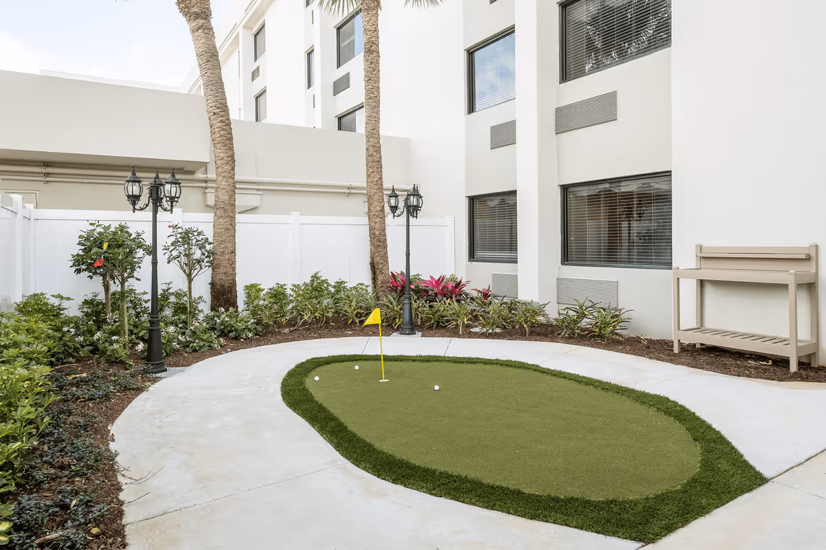 Small outdoor putting green with three golf balls and a yellow flag, surrounded by a concrete walkway, plants, palm trees, and a white fence. The background shows the exterior wall of a building with windows and a beige bench.
