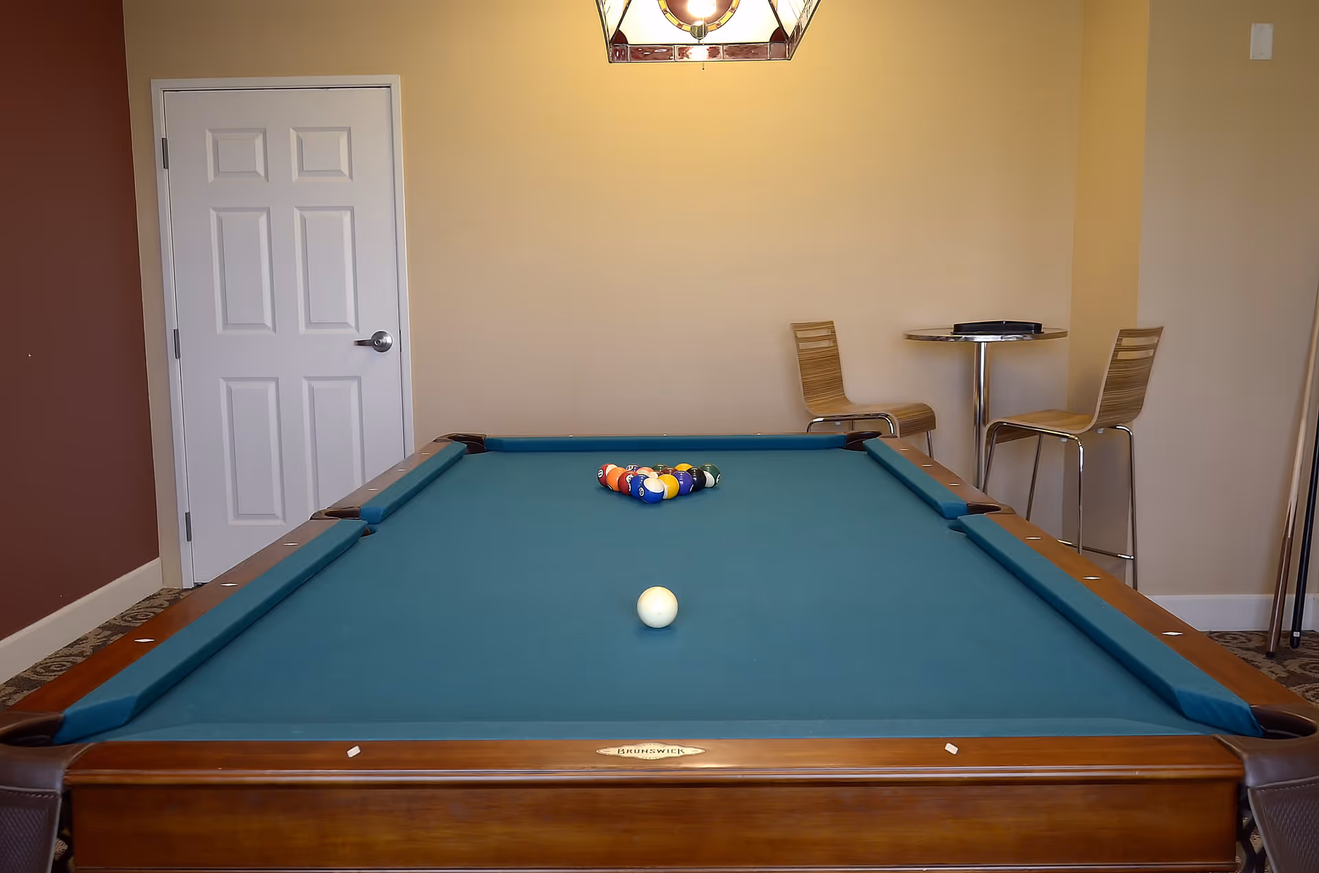 Interior room with a pool table set up for a game, featuring a blue felt surface and a rack of billiard balls. In the background, there is a small round table with two wooden chairs, a closed white door, and a wall-mounted light fixture above.