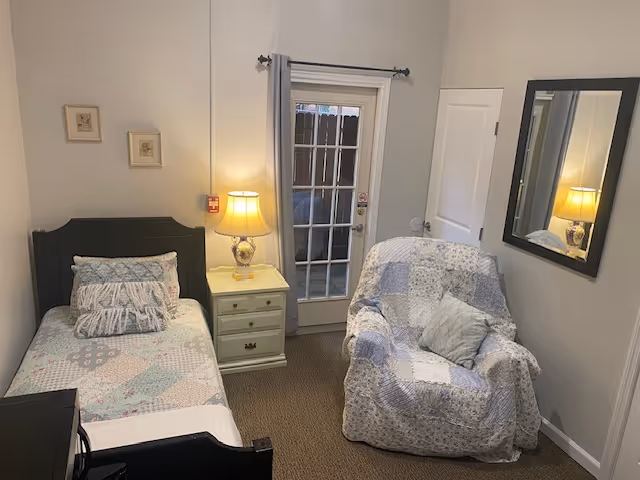 A small, cozy bedroom with a single bed on the left side covered with a pastel patchwork quilt and matching pillows. Next to the bed is a cream-colored nightstand with a decorative lamp. A cushioned armchair with a similar patchwork cover is placed near a glass door with gray curtains. A large mirror hangs on the right wall, reflecting part of the room.