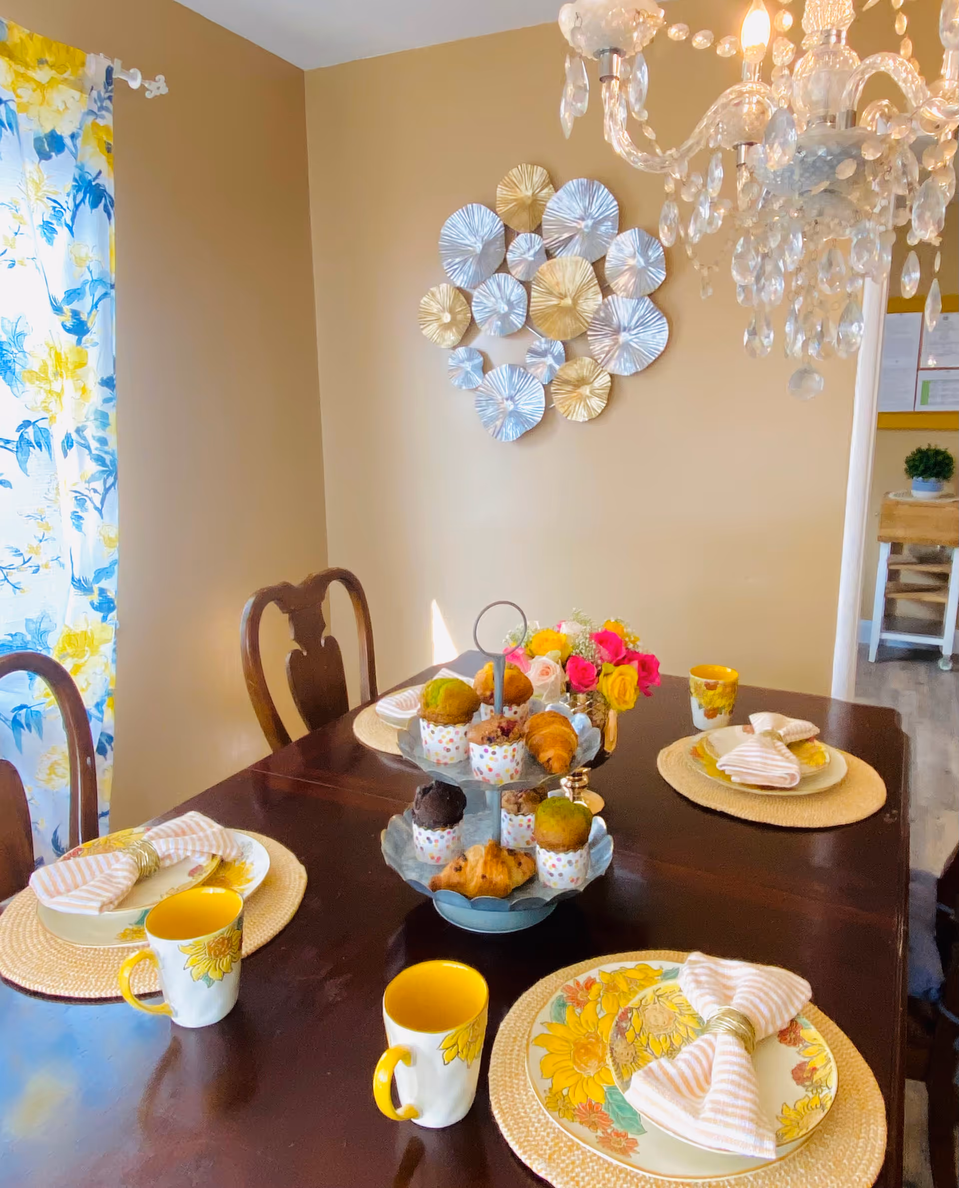 A dining room with a dark wooden table set for four people. The table has floral-patterned plates, striped napkins, sunflower-themed mugs, and a two-tiered tray with assorted muffins and croissants. A colorful bouquet of flowers is also on the table. The room has beige walls, a decorative metal wall art piece, floral curtains, and a crystal chandelier hanging above the table.