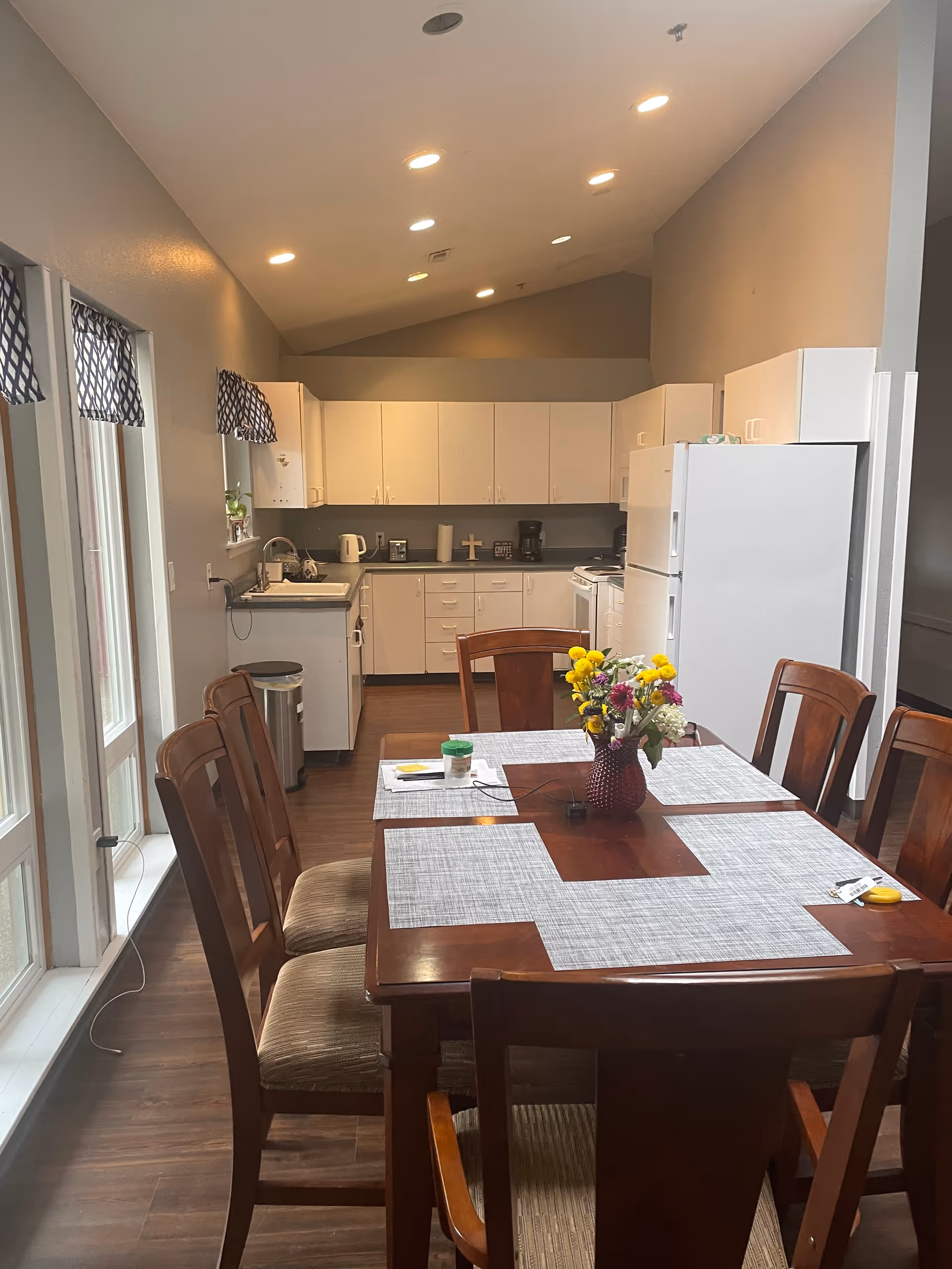 Communal dining area with a wooden table and chairs and a vase of flowers in front of a white kitchen with cabinets and appliances.