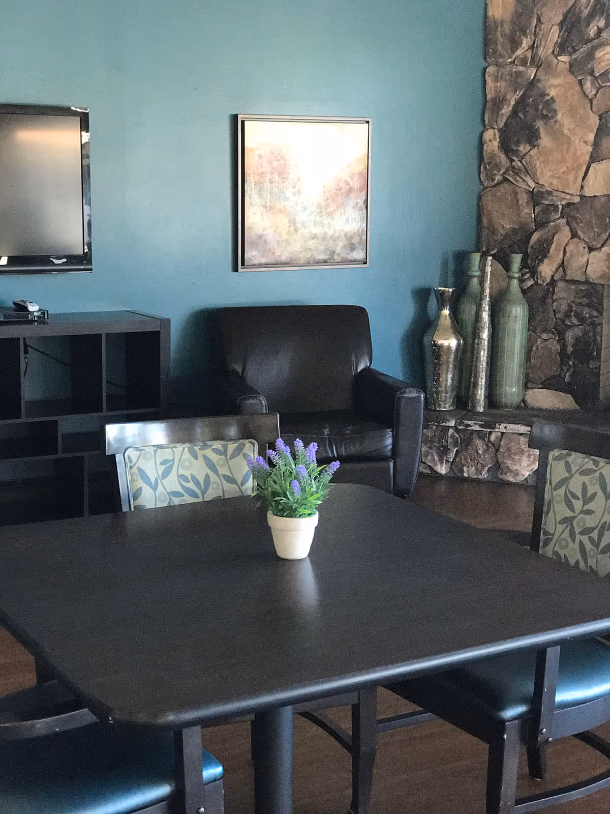 A cozy interior room with a dark wooden table and four chairs, two of which have patterned cushions. A small potted plant with purple flowers is centered on the table. Behind the table is a black leather armchair, a wall-mounted TV, a black shelving unit, a framed artwork on a teal wall, and a stone fireplace with decorative vases.