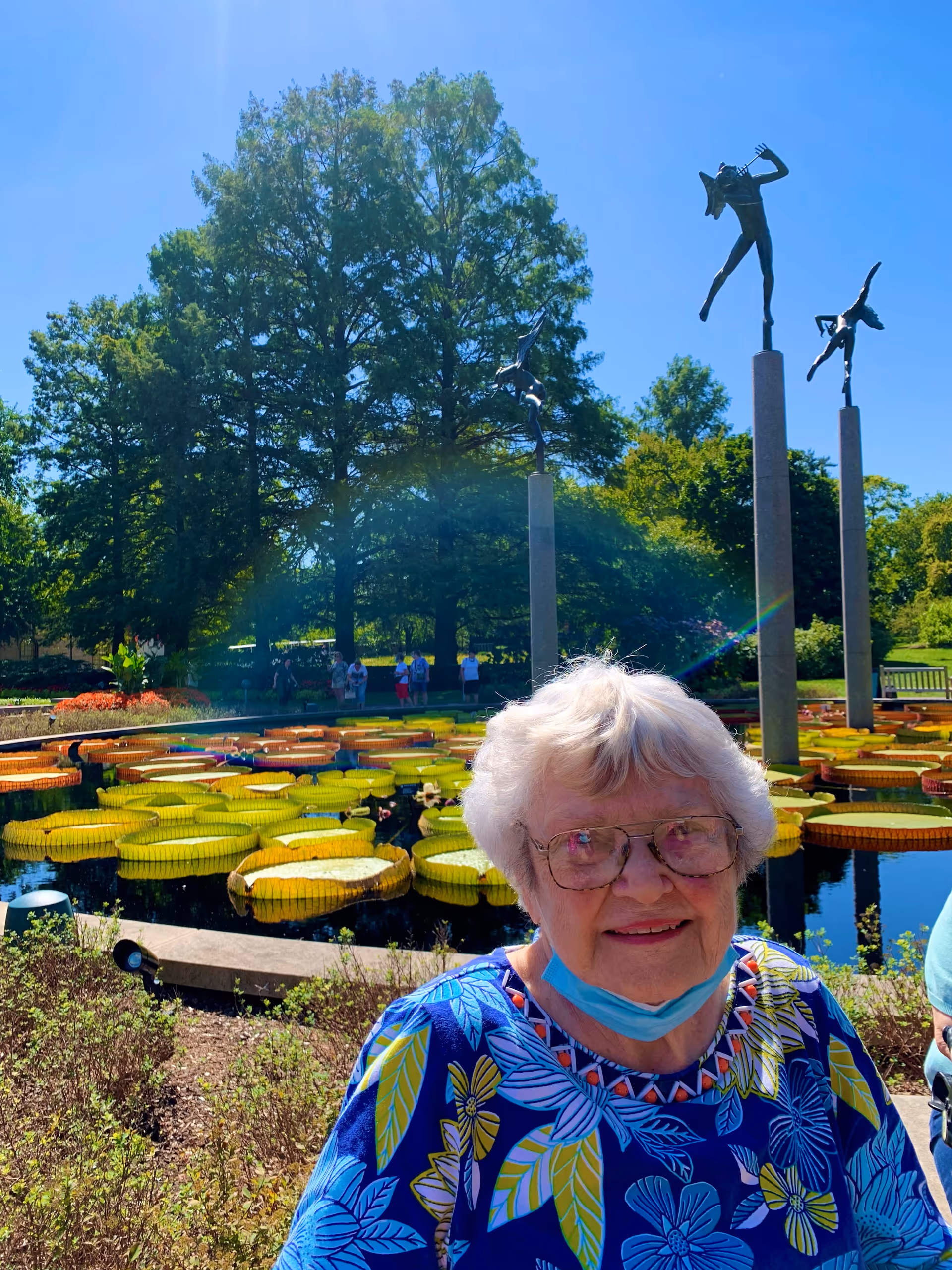 An elderly woman wearing glasses and a blue floral shirt stands outdoors near a pond with large lily pads. Behind her are tall trees, three tall pillars with sculptures of dancing figures on top, and several people in the background enjoying the sunny day.