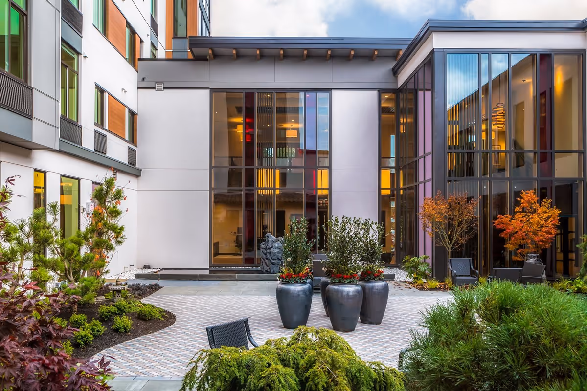 A modern outdoor courtyard at Aegis Gardens Newcastle featuring large windows, potted plants, small trees with autumn foliage, and outdoor seating surrounded by the building's exterior walls.