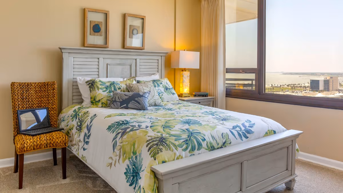 A bright bedroom with a large window showing a city and water view. The room features a white wooden bed with a floral patterned bedspread in green and blue tones, a yellow patterned chair with a laptop on it, a nightstand with a lamp, and two framed pictures on the wall above the bed.