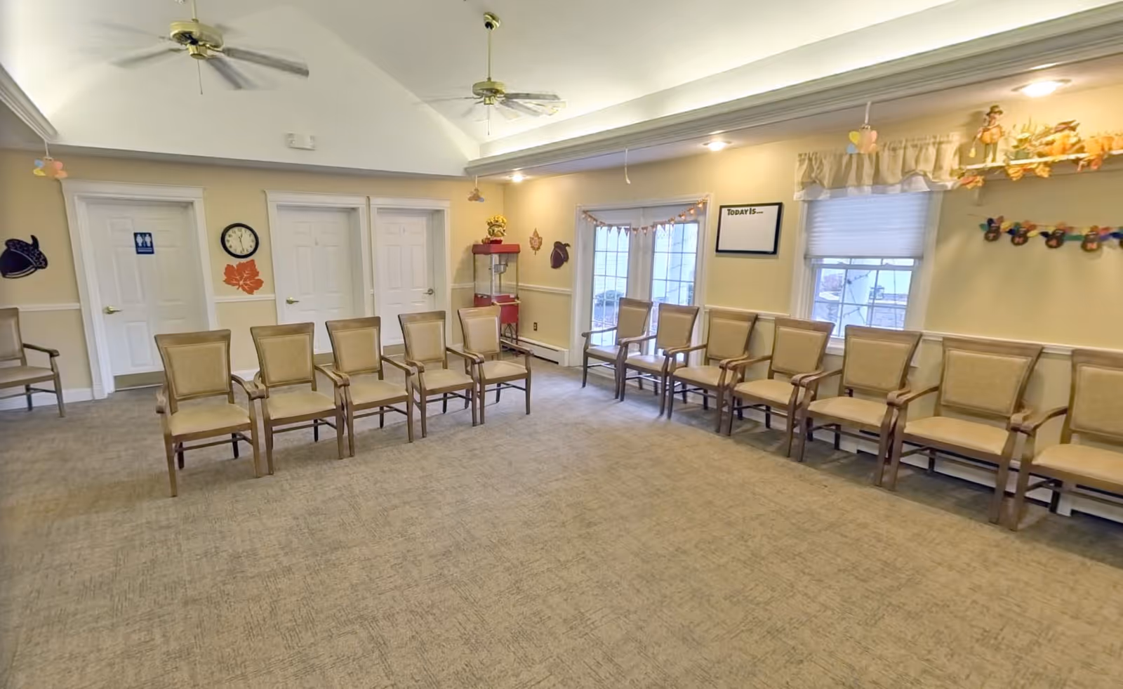 A spacious room with beige carpet and cream-colored walls, featuring two rows of wooden chairs with cushioned seats and backs arranged facing each other. The room has a high ceiling with two ceiling fans, three white doors on one wall, a clock, and decorative wall hangings. There are windows with curtains and a glass door letting in natural light. A popcorn machine is visible in the corner.