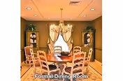 A formal dining room with a round wooden table surrounded by six chairs. The room features a chandelier hanging above the table, two glass-front cabinets with decorative items inside, and a window with curtains in the background. The walls are painted in a warm tone and there are plants on top of the cabinets.