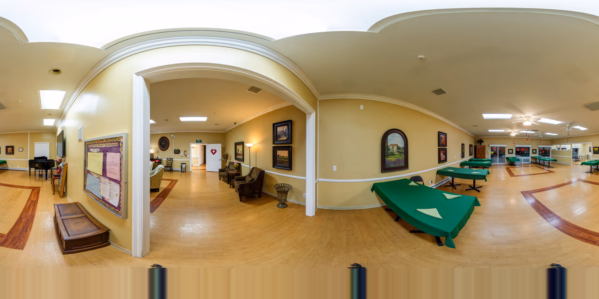 Wide panoramic view of a senior living facility common area with multiple green-covered tables arranged for activities or dining. The room has light yellow walls adorned with framed paintings and a bulletin board. There are several chairs and a small seating area with armchairs and a lamp near an arched doorway. The floor is wooden with darker wood inlays, and the ceiling has recessed lighting and ceiling fans.