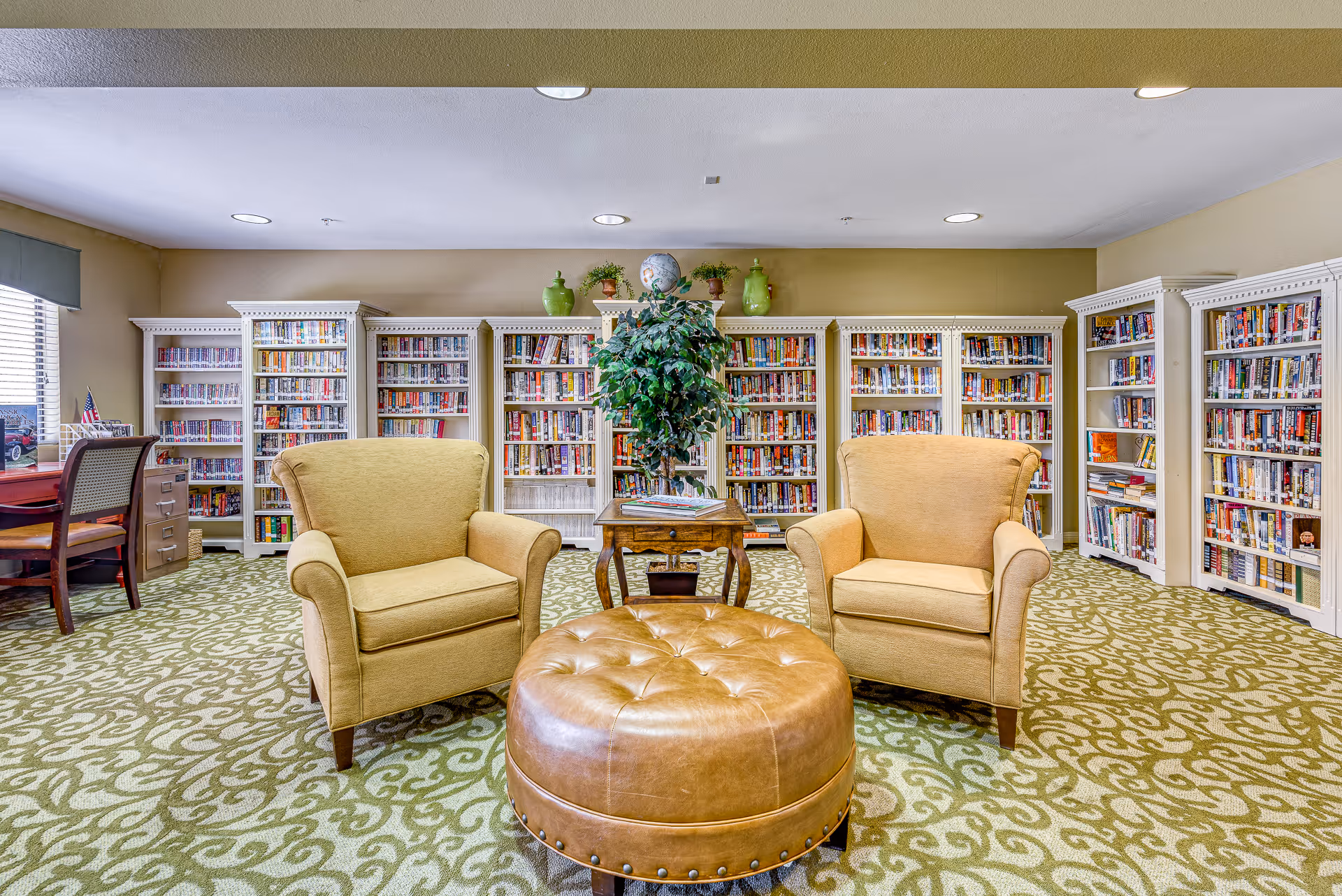 Cozy senior living library with two armchairs, a round leather ottoman, bookshelves lining the back wall, and a small table with a plant.