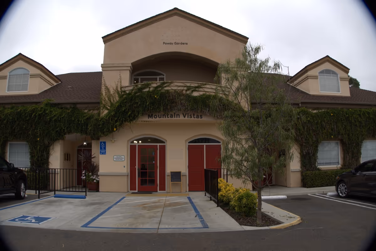 Exterior view of a senior living facility named Poway Gardens with a beige building facade covered partially in green ivy. The entrance has two red doors under an archway labeled Mountain Vistas. There are handicap parking spaces and a small tree with shrubs in front of the building.