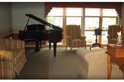 A cozy living room area with a grand piano near large windows. The room features striped armchairs, a white sofa, a wooden side table with a lamp, and a small round table with a plant. Natural light fills the space through the windows.