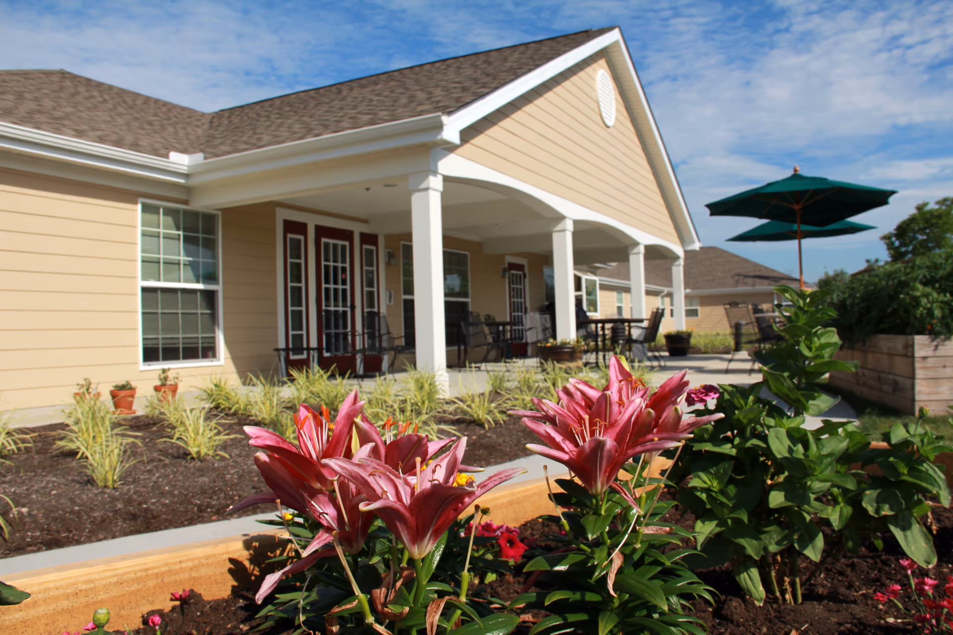 Outdoor garden area with blooming pink lilies in the foreground and a beige senior living facility building with a covered porch, patio furniture, and green umbrellas under a blue sky in the background.