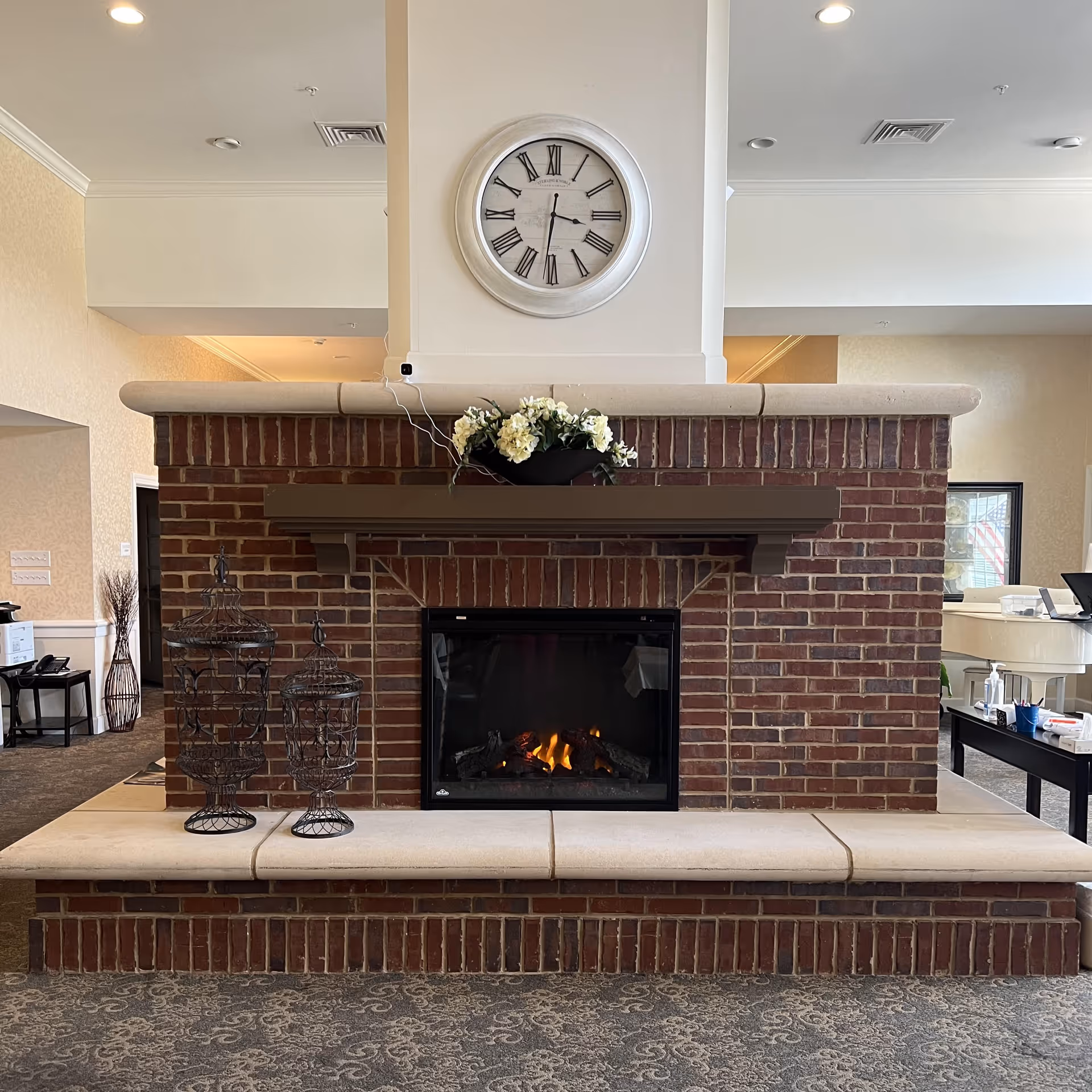 Interior view of a common area in an assisted living facility featuring a brick fireplace with a fire burning inside. Above the fireplace is a large round clock with Roman numerals. On the mantel, there is a flower arrangement. To the left of the fireplace are two decorative metal lanterns. The room has carpeted flooring and light-colored walls with recessed ceiling lights.